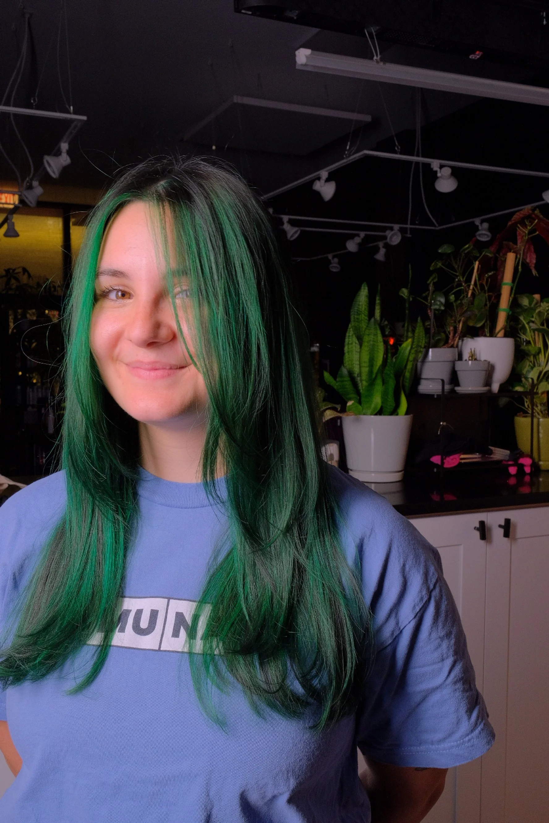 A woman with long black hair that has green streaks, wearing a blue t-shirt with a printed design, smiling and standing indoors near a counter with potted plants.