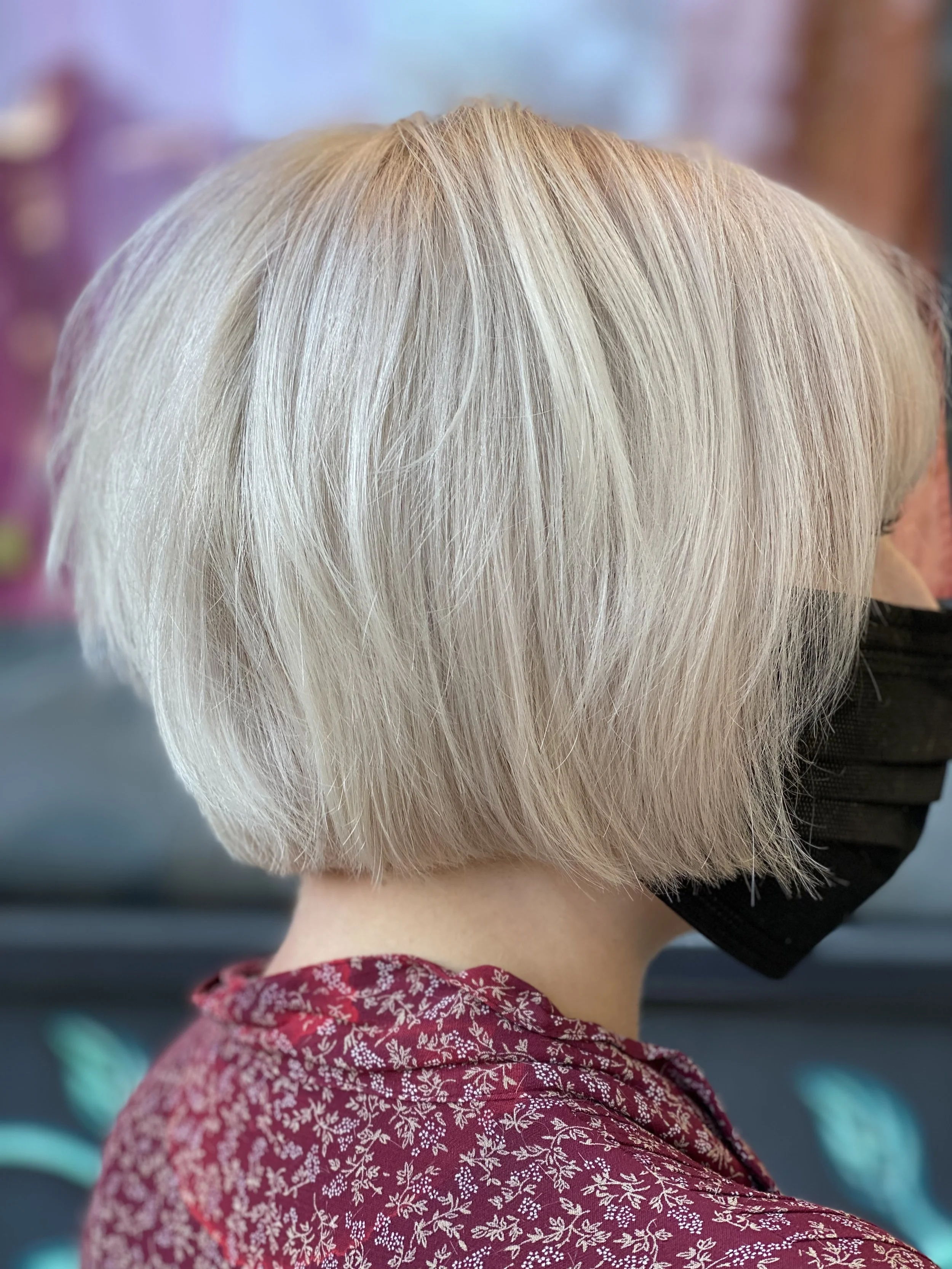 Back view of a person with short, platinum blonde hair wearing a black face mask and a maroon shirt with white floral pattern.