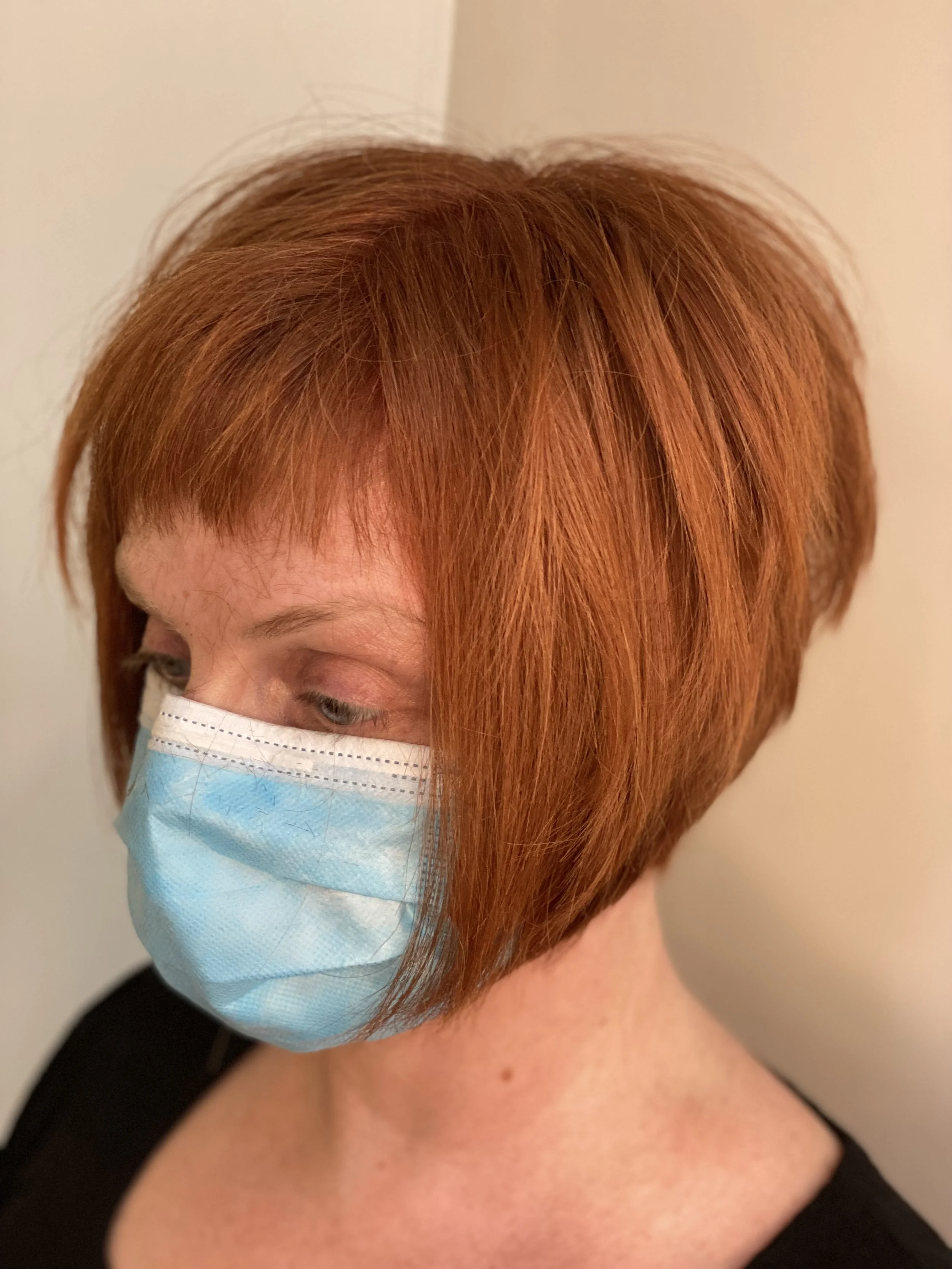 A woman with short red hair wearing a light blue face mask, turned slightly to the side, against a plain background.