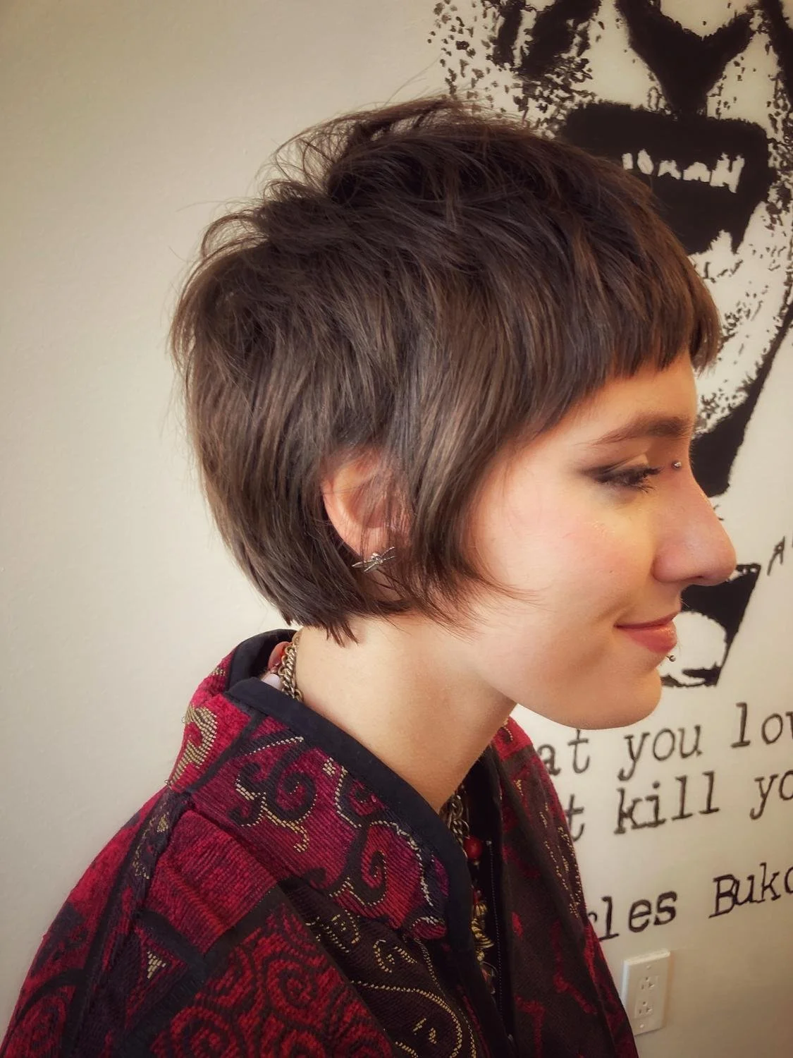 Side profile of a woman with short, layered brown hair, wearing earrings and a red and black patterned jacket, standing in front of a wall with black and white artwork and text.