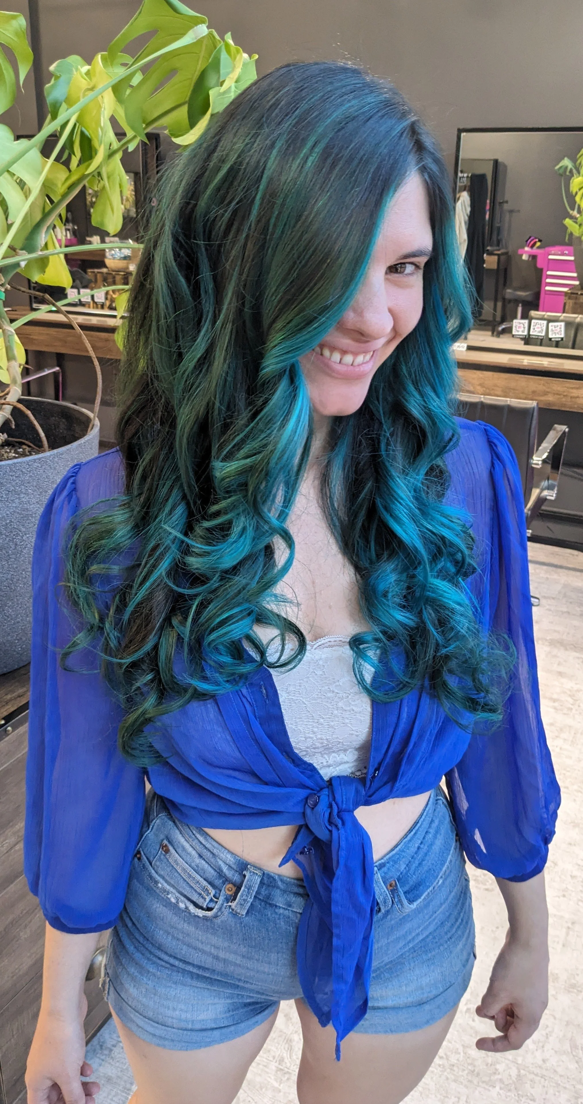 Young woman with long, curly hair dyed in shades of blue and green, smiling, wearing a blue tied-up blouse and denim shorts, standing in a salon with plants and mirrors in the background.