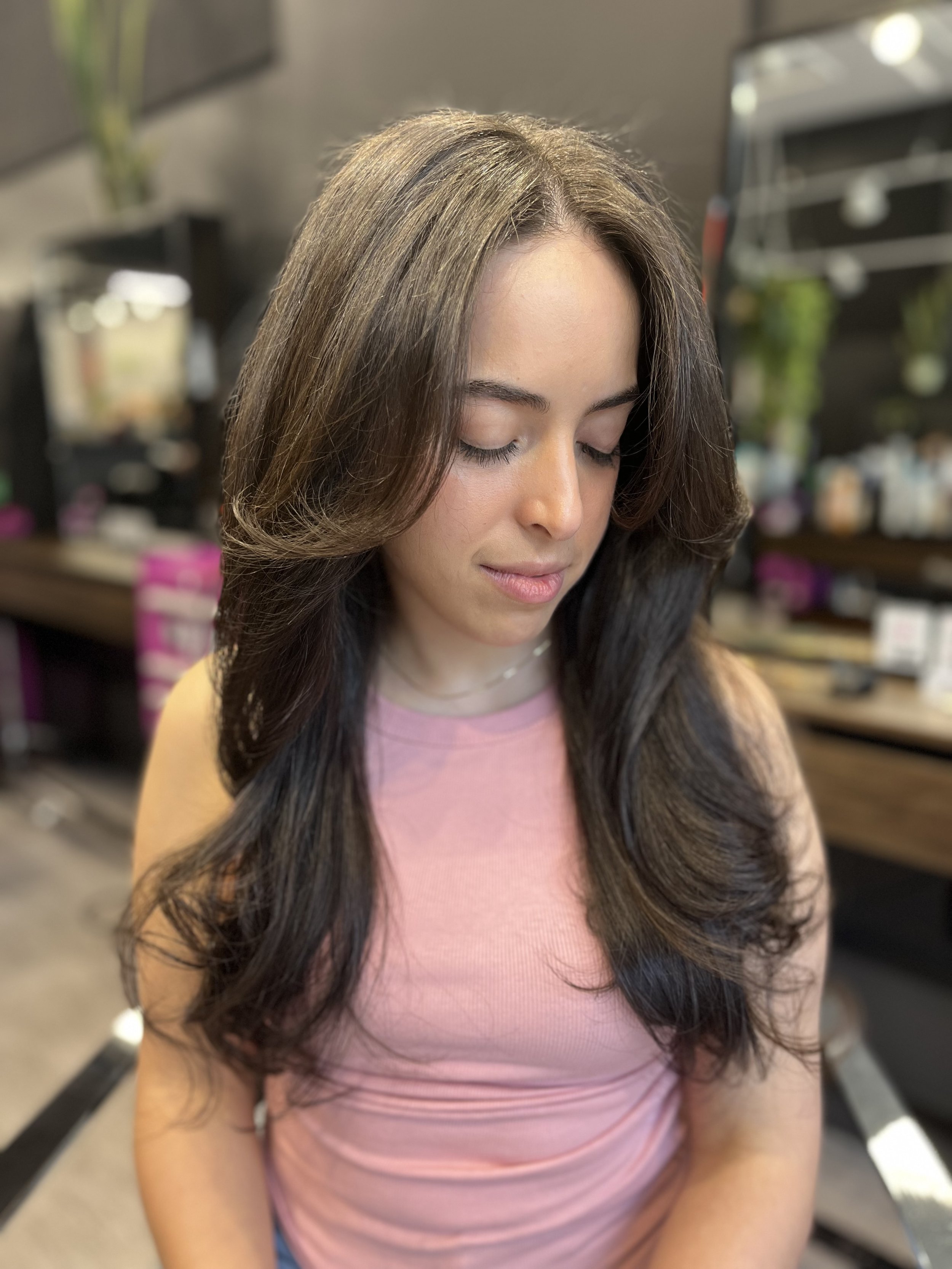 A young woman with long dark hair and light skin, wearing a pink sleeveless top and a delicate necklace, sitting in a salon or hair studio, with a blurred background of salon equipment and plants.
