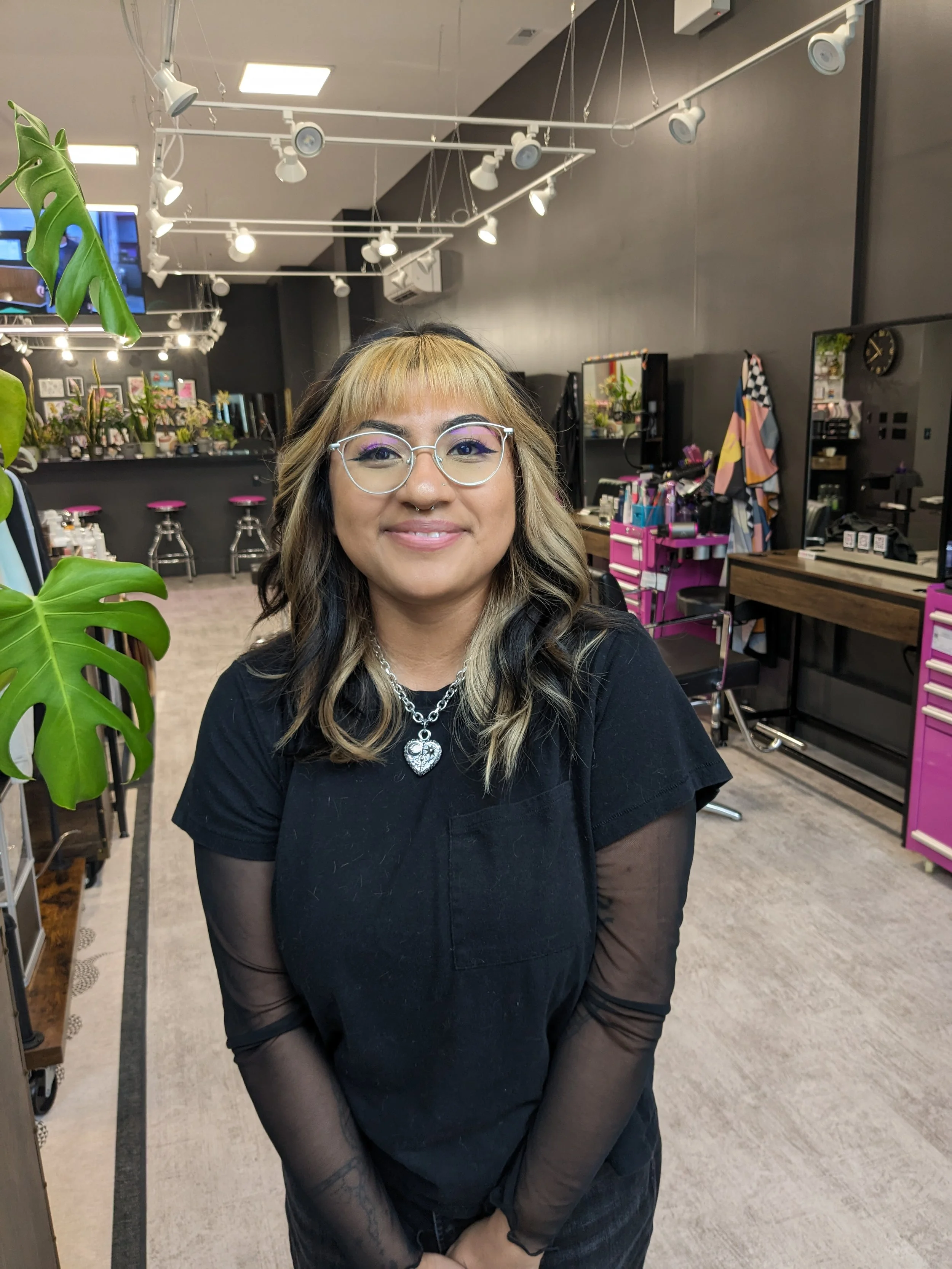 A woman with shoulder-length, wavy black and blonde hair, wearing glasses, a black shirt with sheer long sleeves, and a heart-shaped necklace, standing inside a modern salon with black walls, pink cabinets, and styling stations.