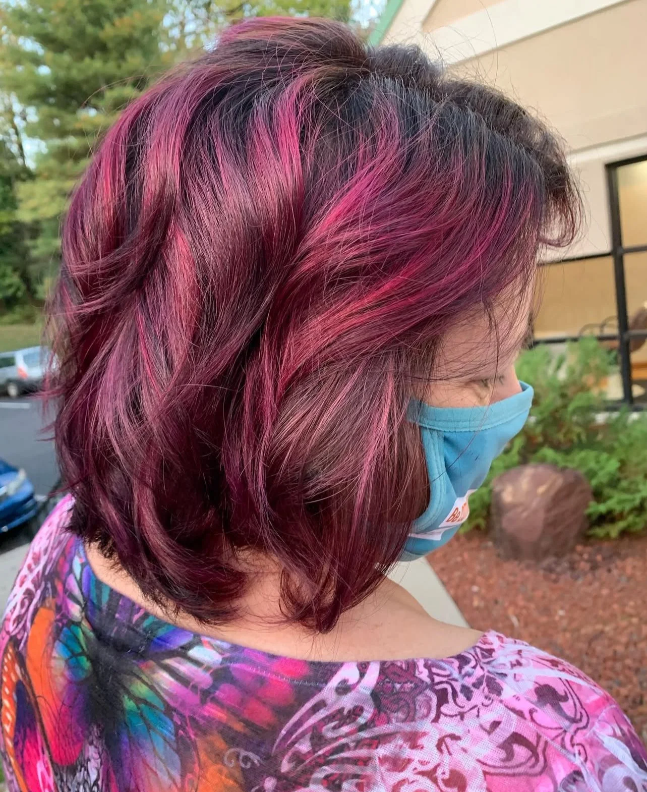 Woman with medium-length, wavy hair dyed in shades of pink and purple, wearing a blue face mask and a colorful patterned shirt, outside near a building.