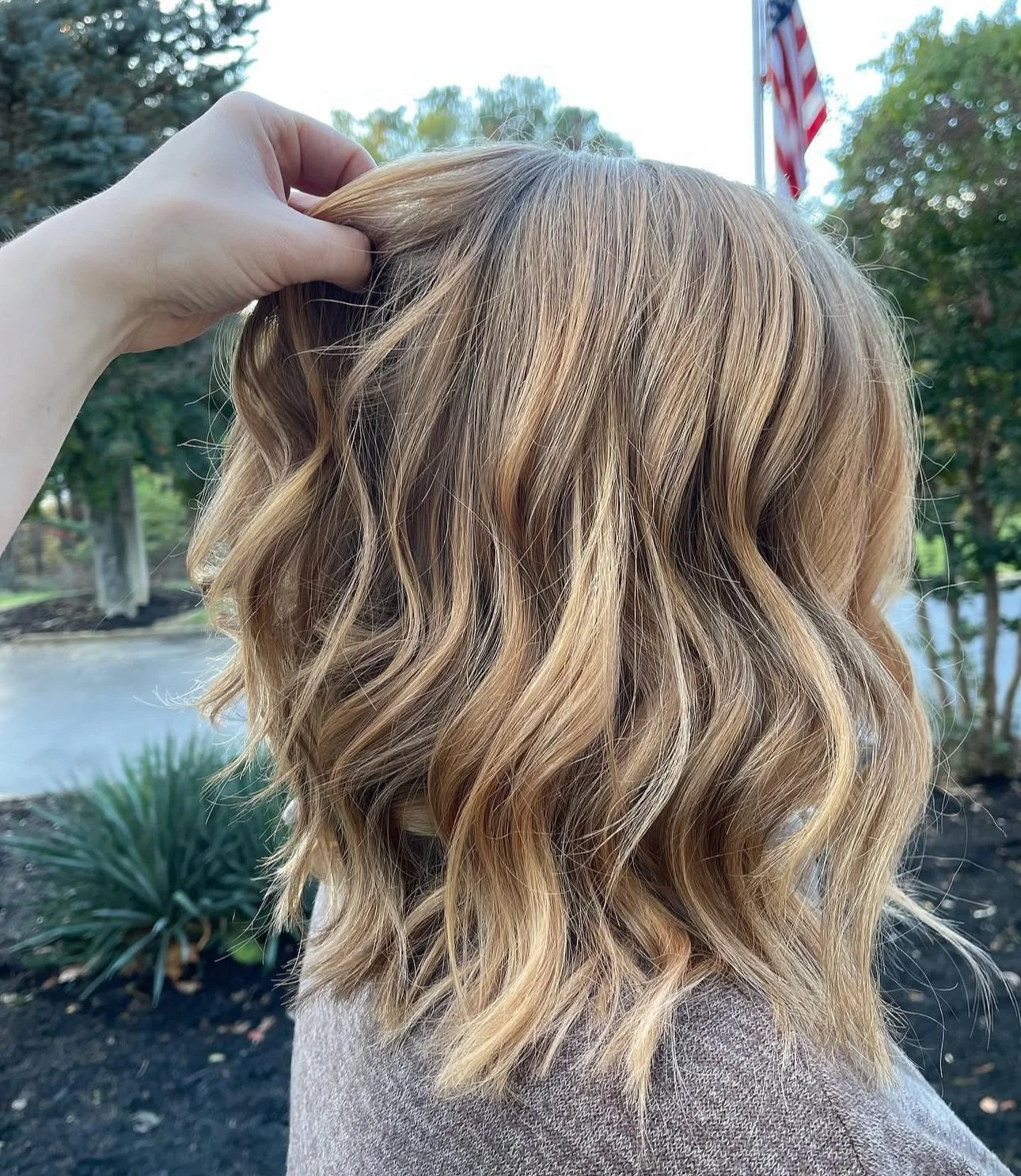 A woman with blonde, wavy hair holding a section of her hair with her hand outdoors during daytime. Trees and an American flag are visible in the background.
