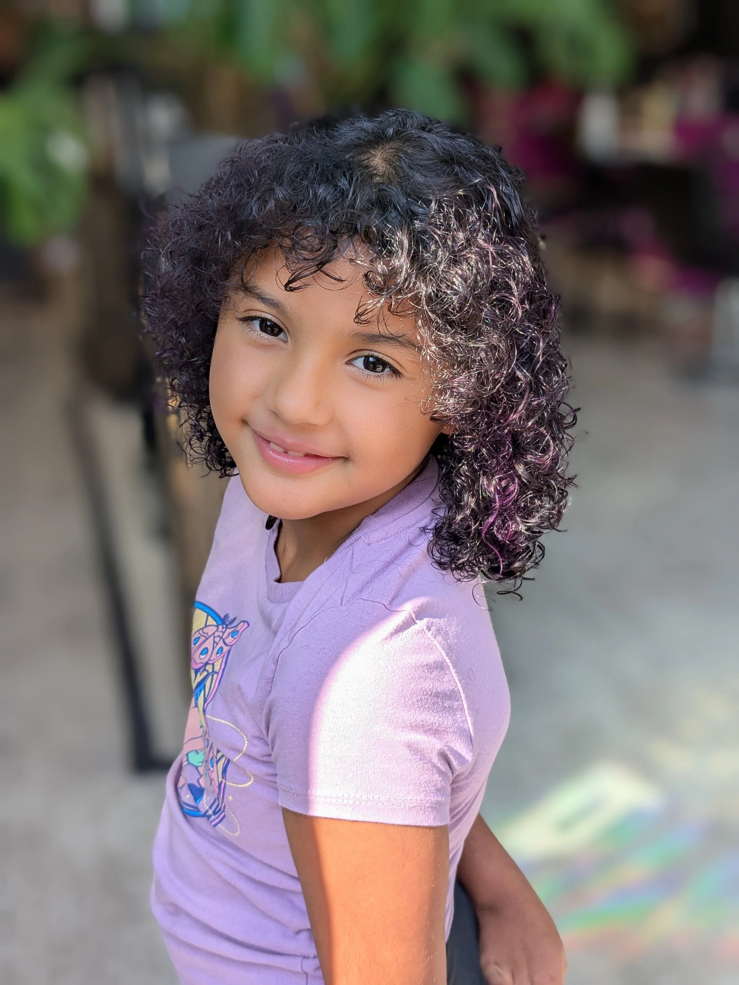 A young girl with curly dark hair and a light purple t-shirt smiling at the camera.