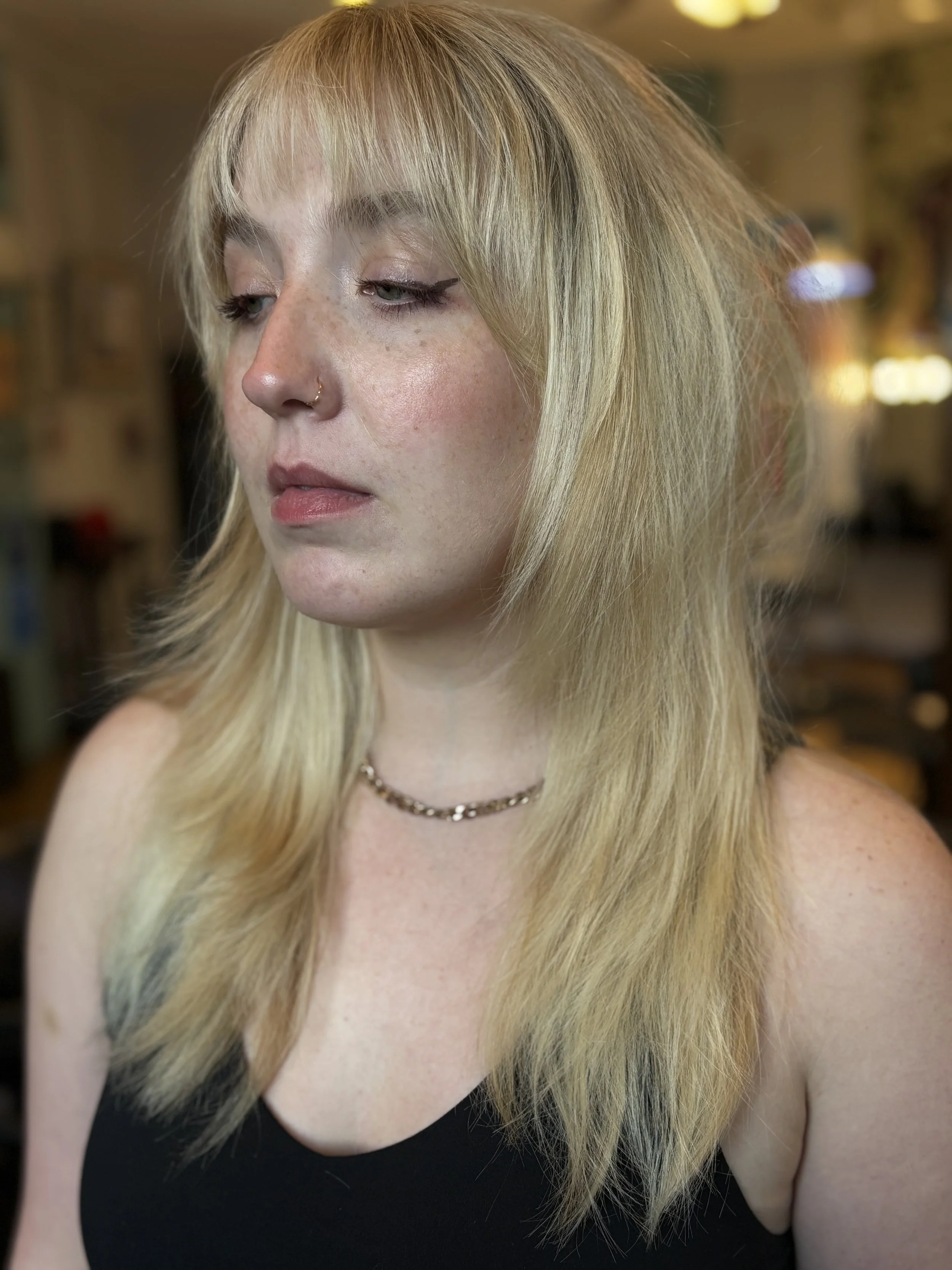 A woman with long blonde hair, light skin with freckles, wearing a black sleeveless top, a nose ring, and a necklace, looking down with a neutral expression. The background is blurred with warm indoor lighting.
