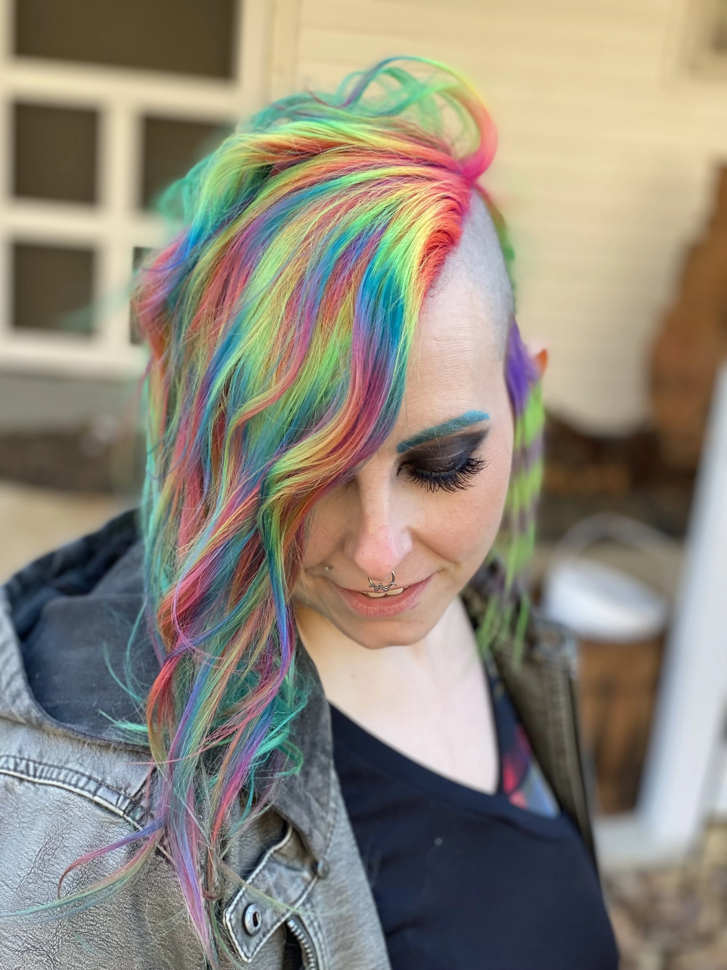 A woman with multi-colored rainbow hair styled in loose waves, wearing dark eye makeup, a septum piercing, and a casual jacket, smiling with her head tilted down.