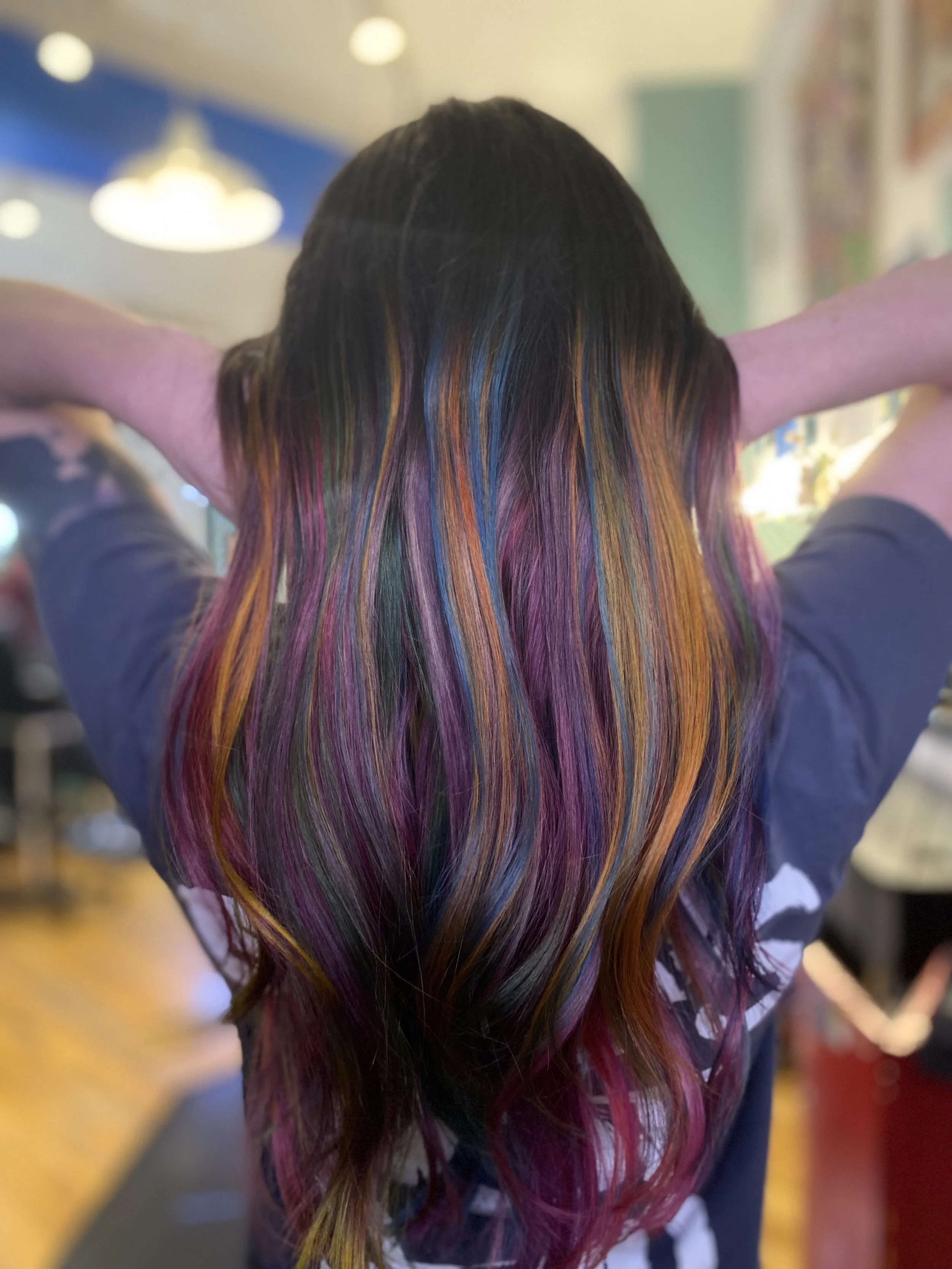 Person with long, wavy hair dyed in multiple vibrant colors including pink, purple, blue, orange, and green, seen from the back.