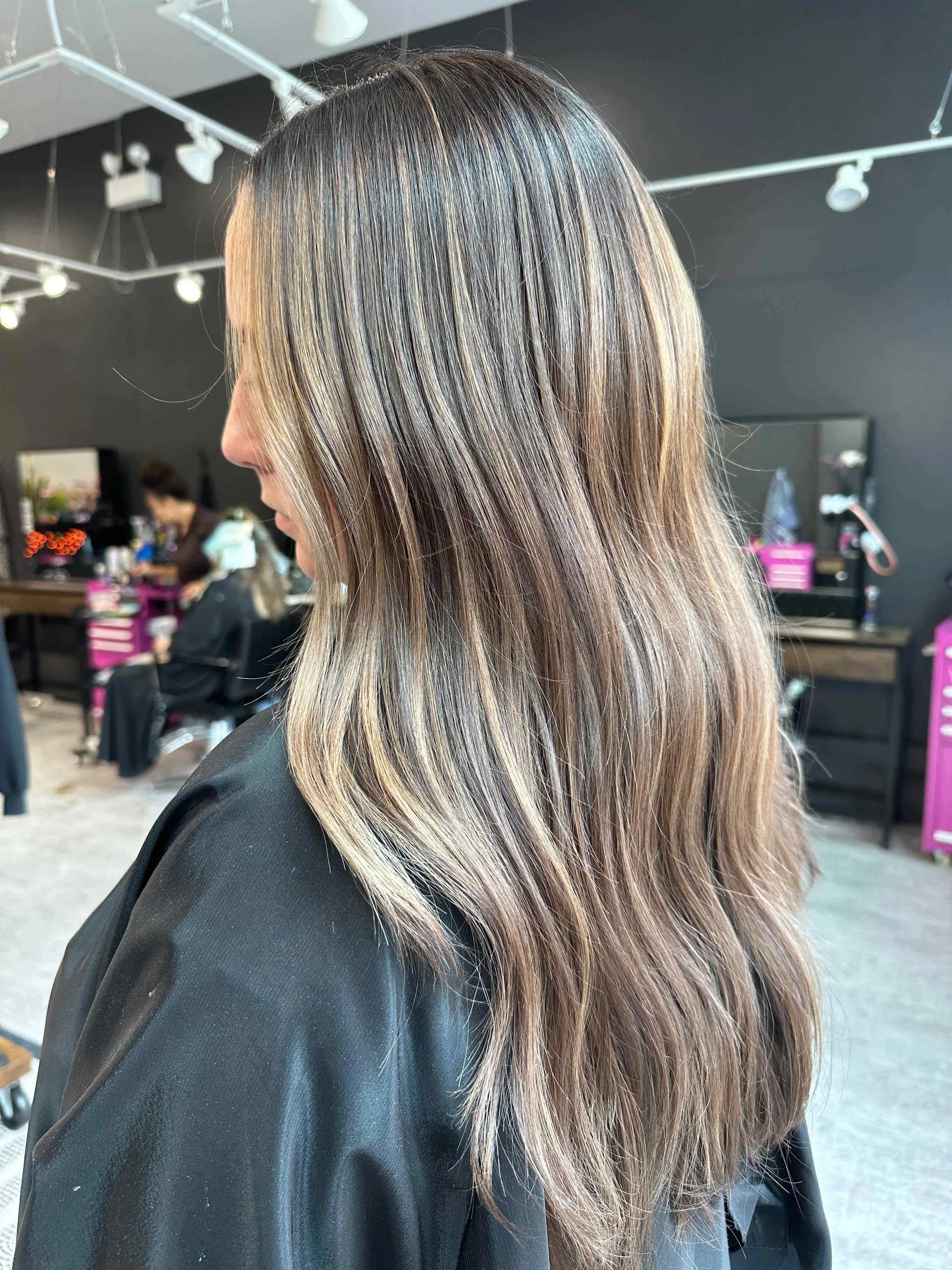 Side view of a woman with long, layered, light brown hair with blonde highlights, sitting in a salon with hairstyling stations and a mirror in the background.