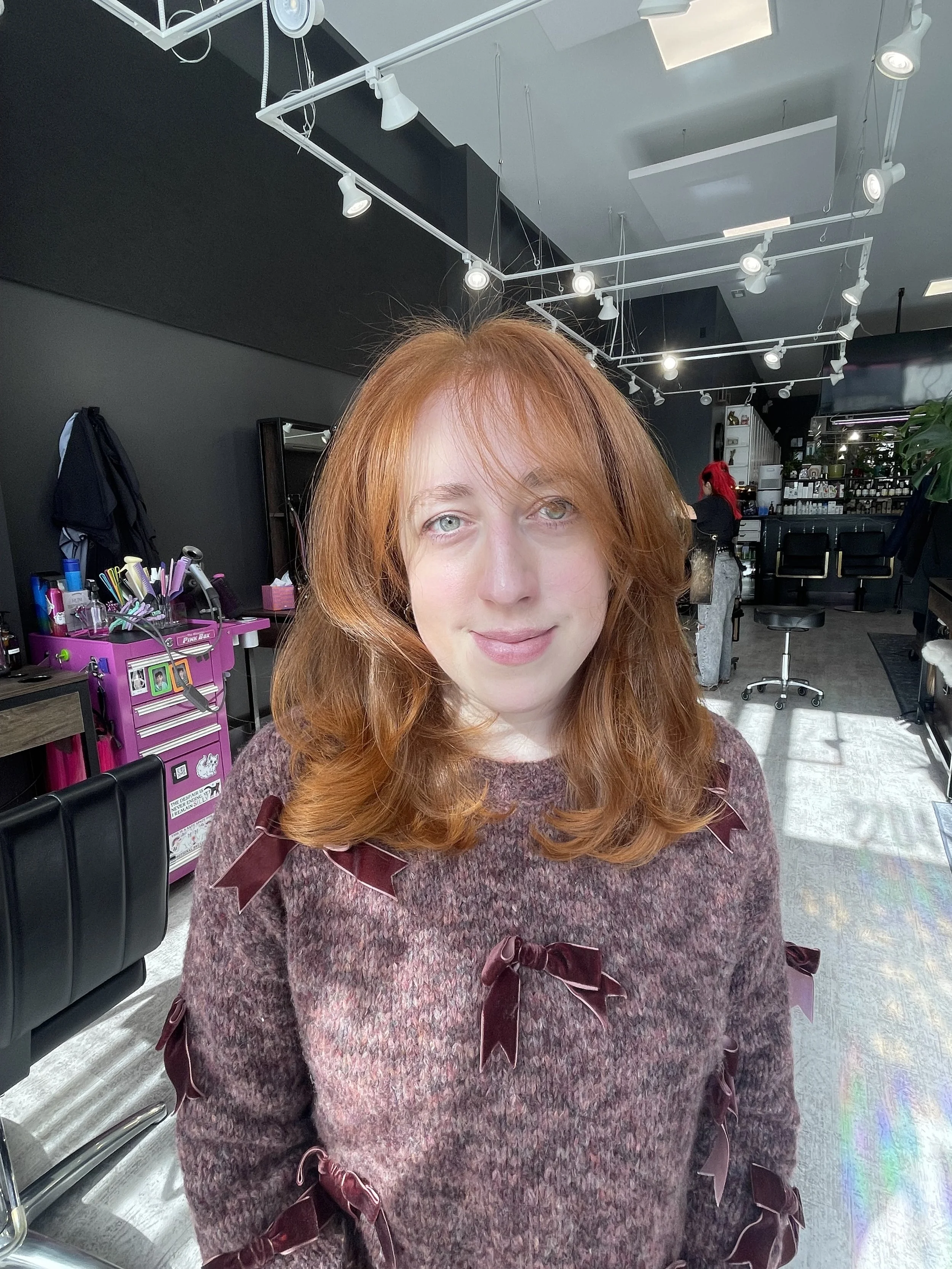 A woman with long, wavy, platinum blonde hair sitting on a green velvet couch in a dimly lit room with plants and a neon sign that says 'Have a great' in pink.