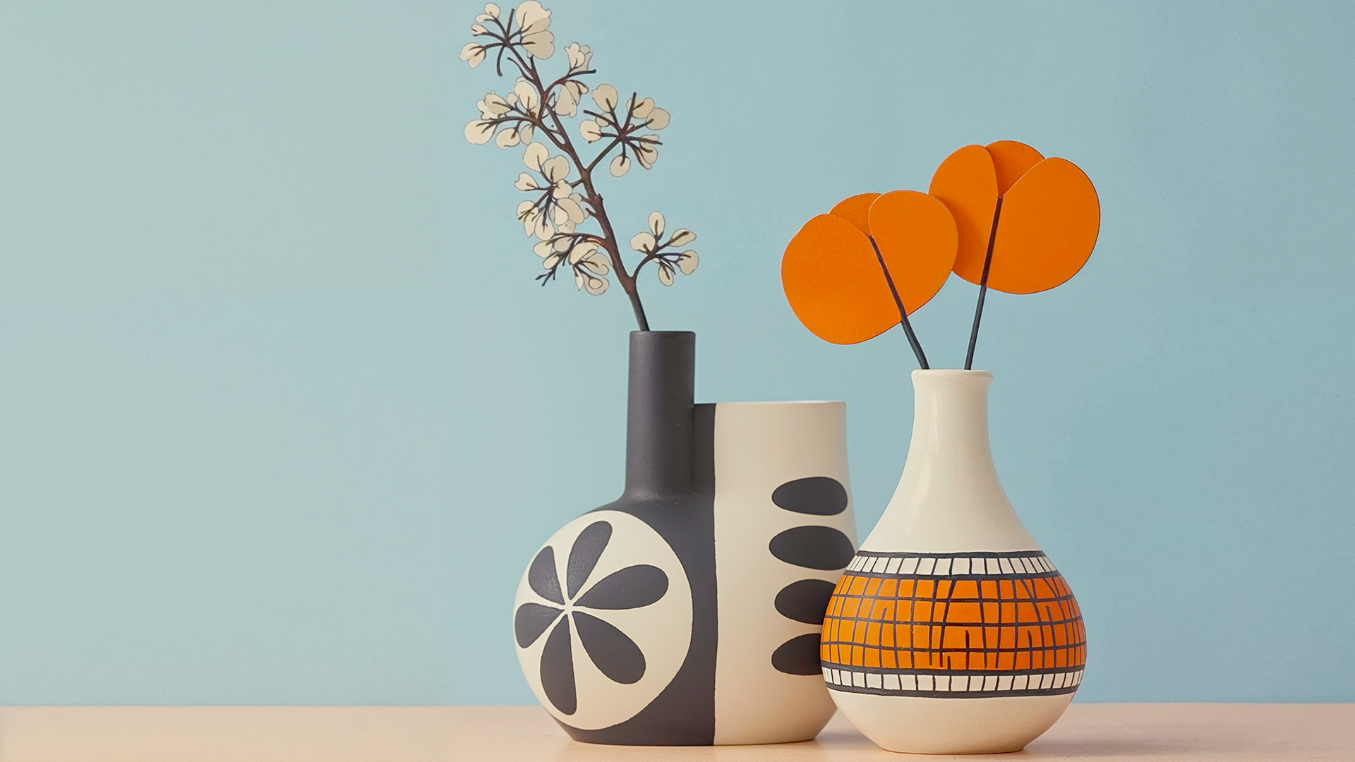 Deux vases décoratifs avec des fleurs en papier, un vase blanc et orange avec des motifs géométriques, un autre vase noir et blanc avec des dessins floraux, placés contre un mur bleu clair.