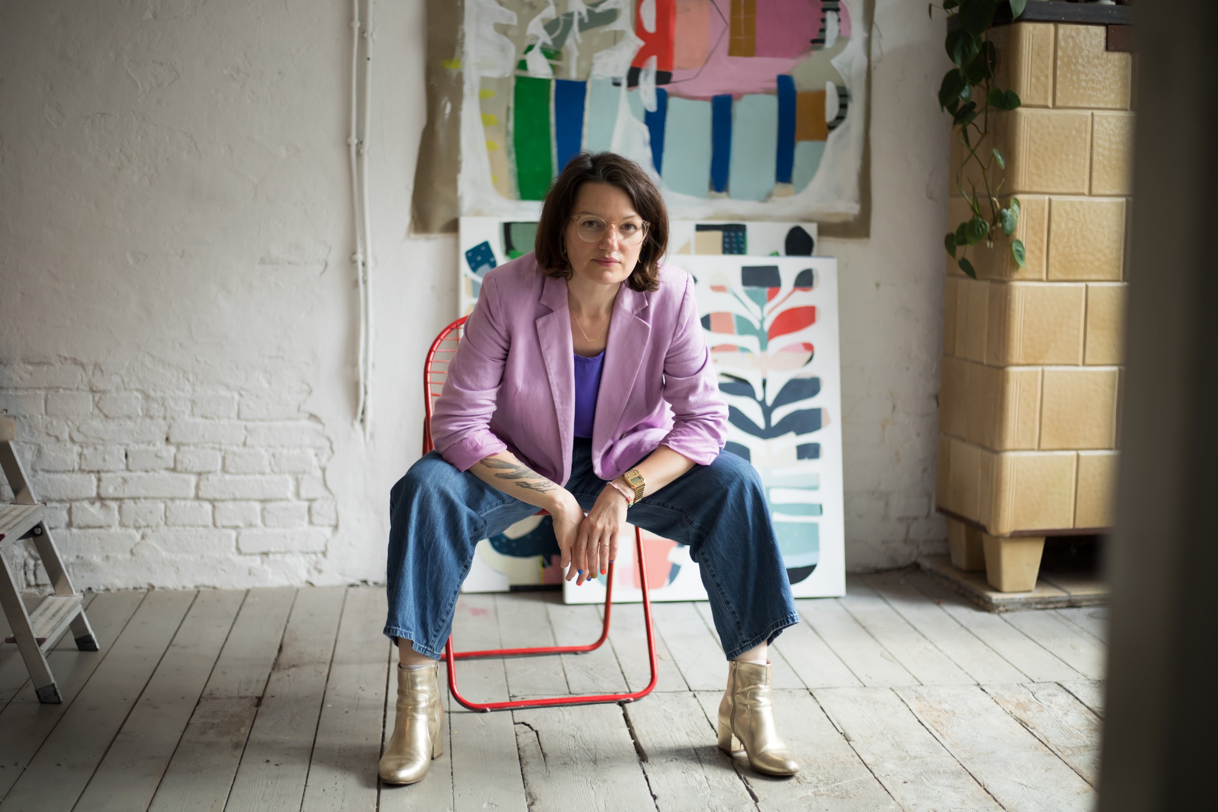 Woman sitting on a red chair in front of an artistic background with colorful paintings, wearing a pink blazer, blue shirt, jeans, and gold ankle boots, with a tattoo on her left forearm and glasses.
