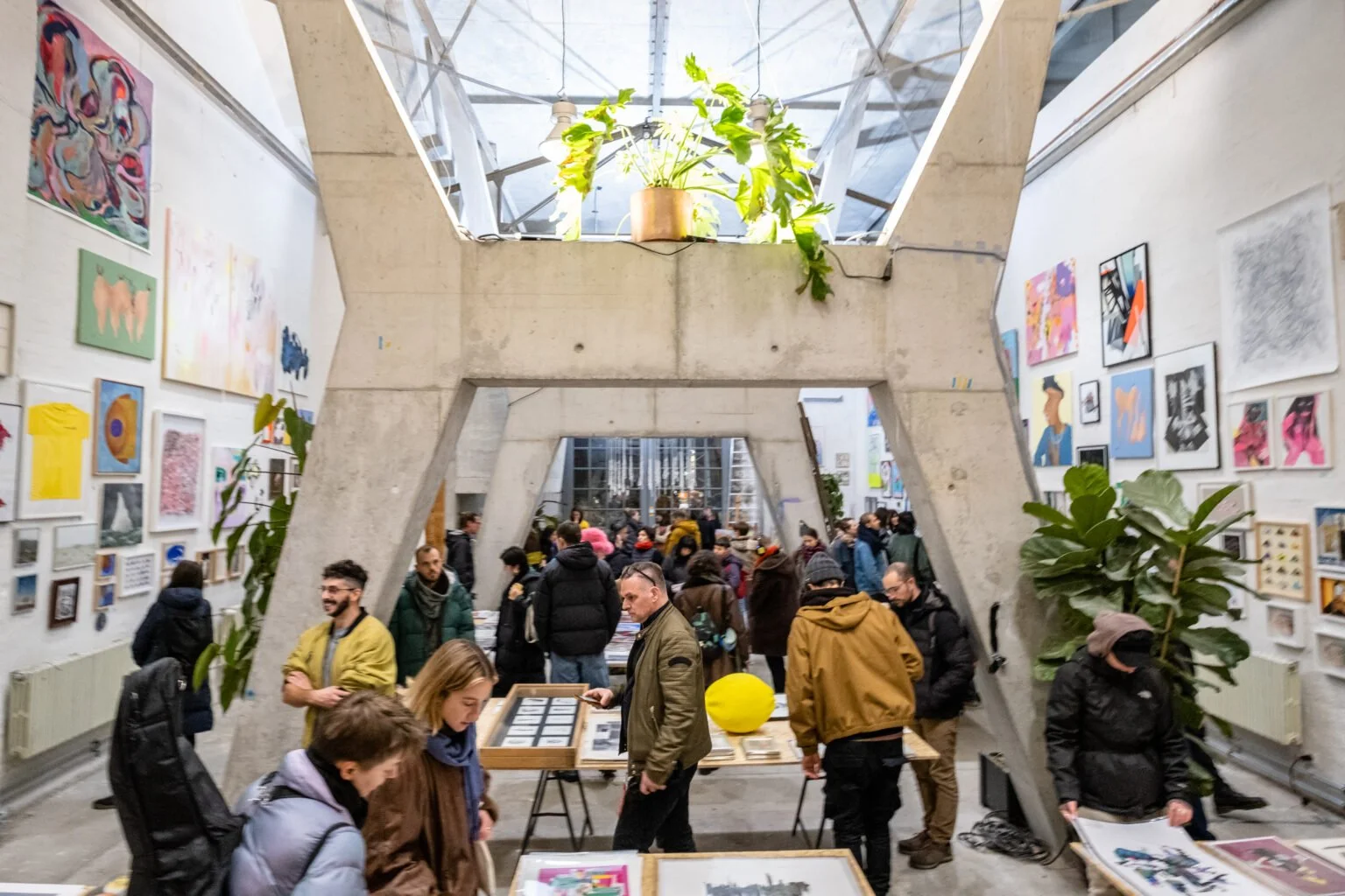 People visiting an art gallery with colorful artwork on the walls, concrete structural beams, and tables displaying art pieces, inside a spacious, industrial-style building.
