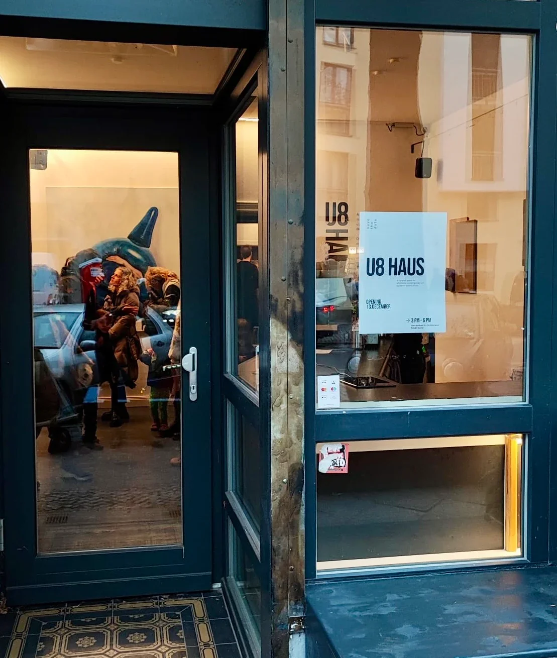 The entrance to U8 Haus, a gallery or exhibition space with a sign indicating opening on December 13th from 3 to 6 PM. The glass door reflects a street scene with people and parked cars.