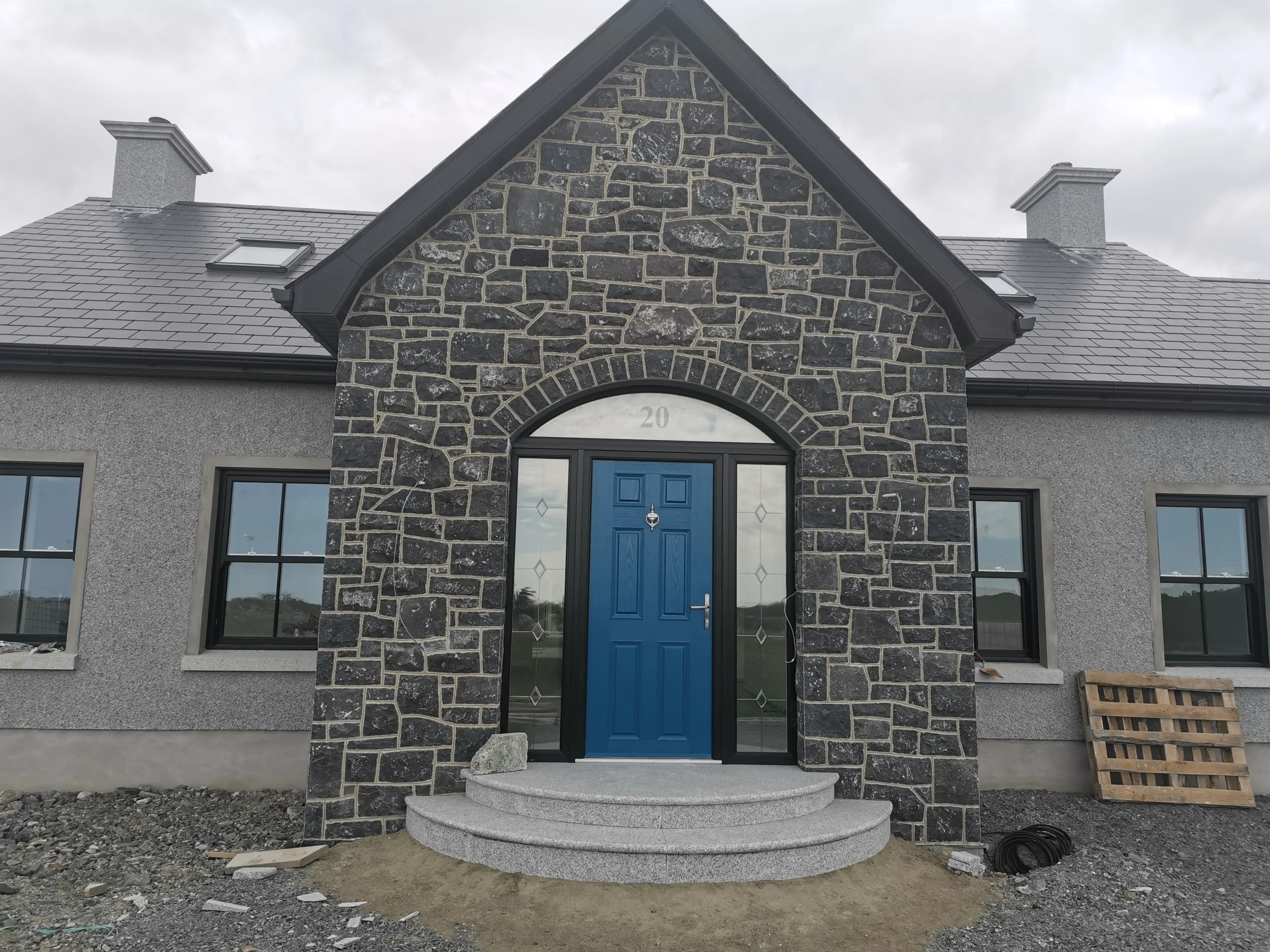 Image shows a porch built with random blue limestone building stone. The porch has grey granite steps and a blue door and the dash is a grey granite dash with black windows