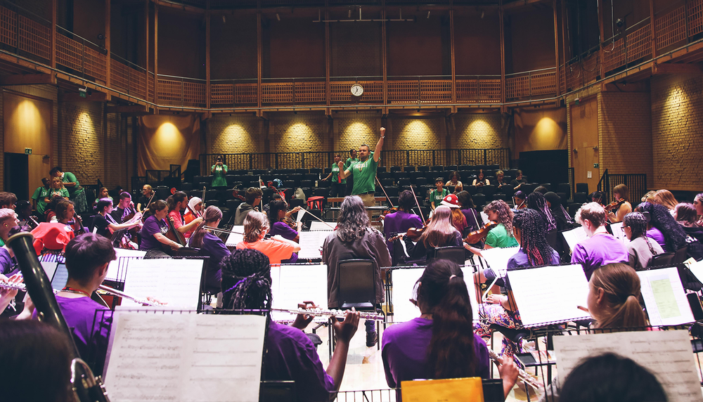 Orchestra practice in a concert hall with a conductor, musicians playing string instruments, and sheet music on stands.