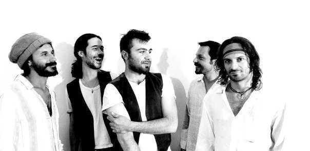 A music group of five men standing together, some smiling and looking at each other, in a black and white photo.