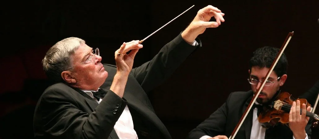 An orchestra conductor and a violinist performing on stage, with the conductor guiding the musicians.