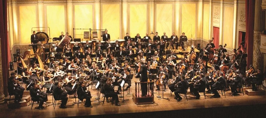 Orchestra performing on stage in a concert hall with a conductor leading them.