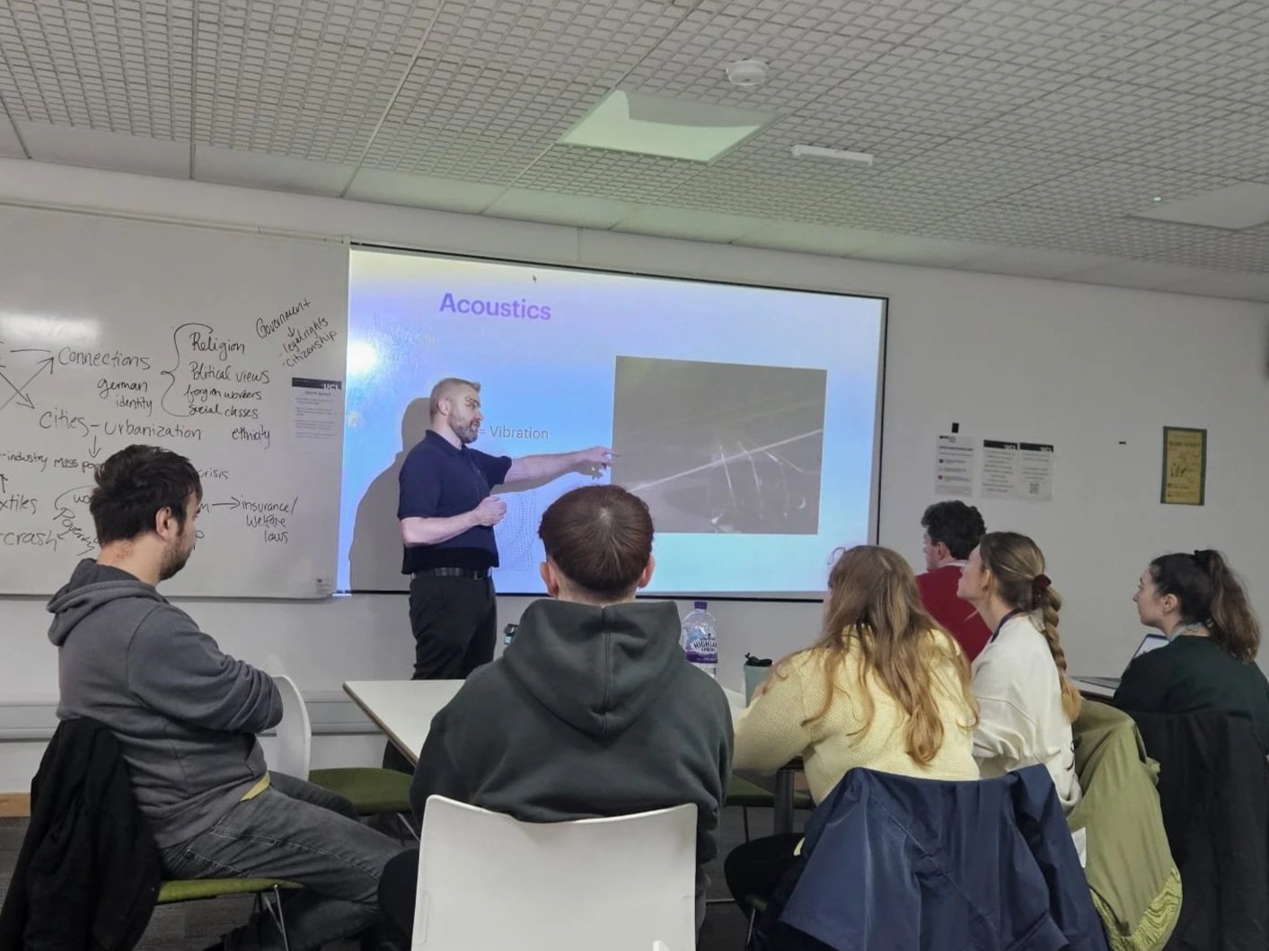 A classroom lecture with a male instructor pointing at a presentation slide titled "Acoustics" on a white screen. Several students are seated at tables, watching and listening, with notes and water bottles on the tables. The classroom has a whiteboard with handwritten notes on the left and framed documents or posters on the wall to the right.