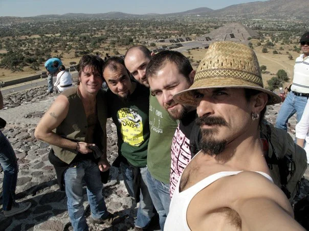 A music group of five men taking a selfie on a rocky archaeological site with hills, sunny and clear weather.