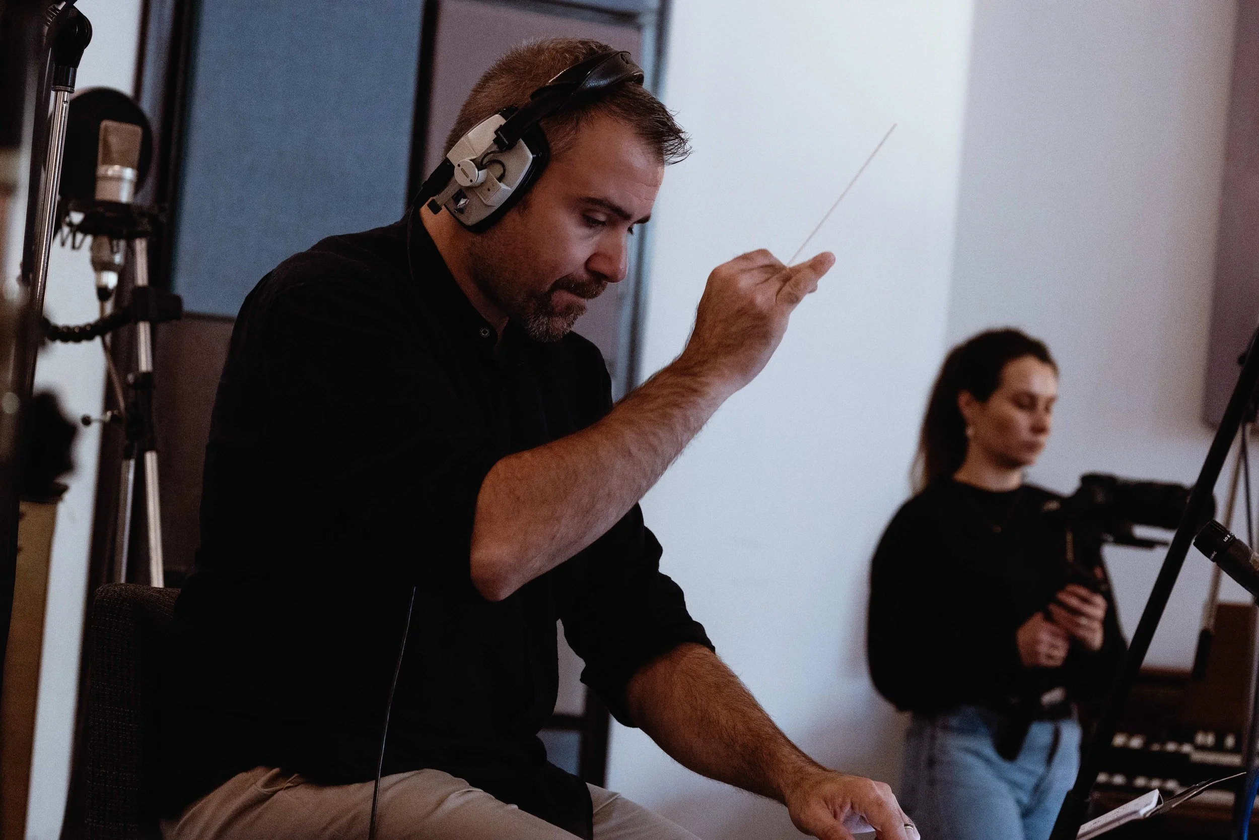 A man in headphones conducts an orchestra with a baton, while a woman in the background takes pictures.
