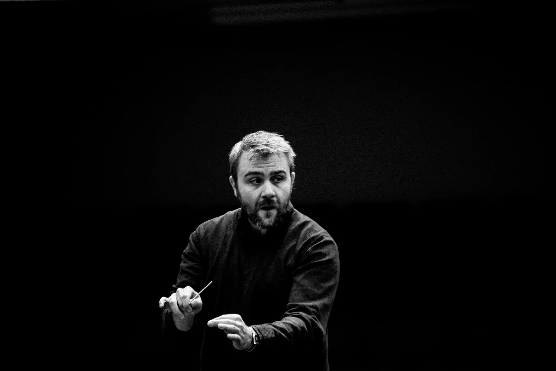 A conductor with a beard and short hair, wearing a dark shirt, holds a baton in one hand and gestures with the other, standing against a dark background.