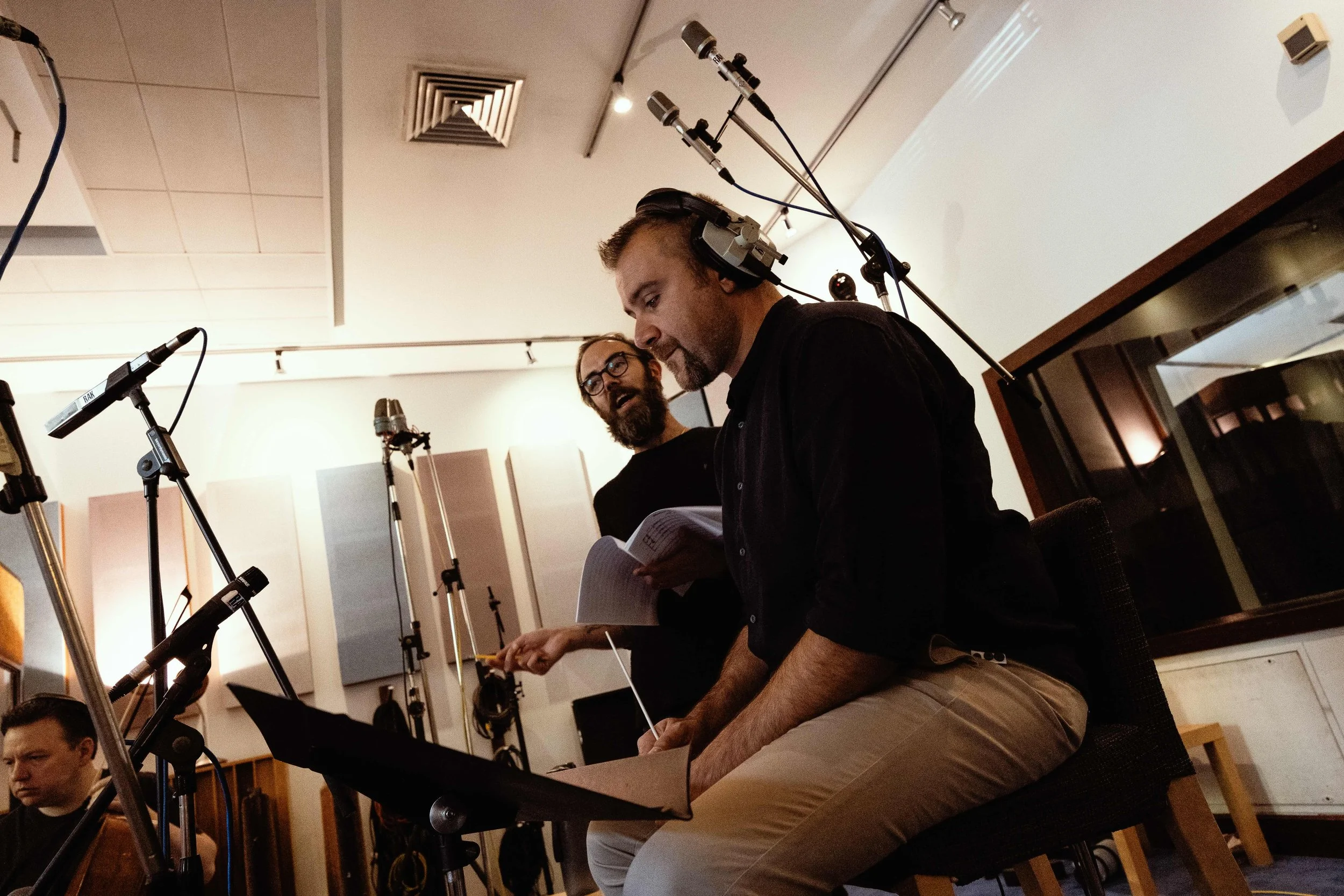 Two men in a recording studio, one with headphones holding a script and point at something, the other with glasses and beard looking at the man with headphones, surrounded by microphones and recording equipment.