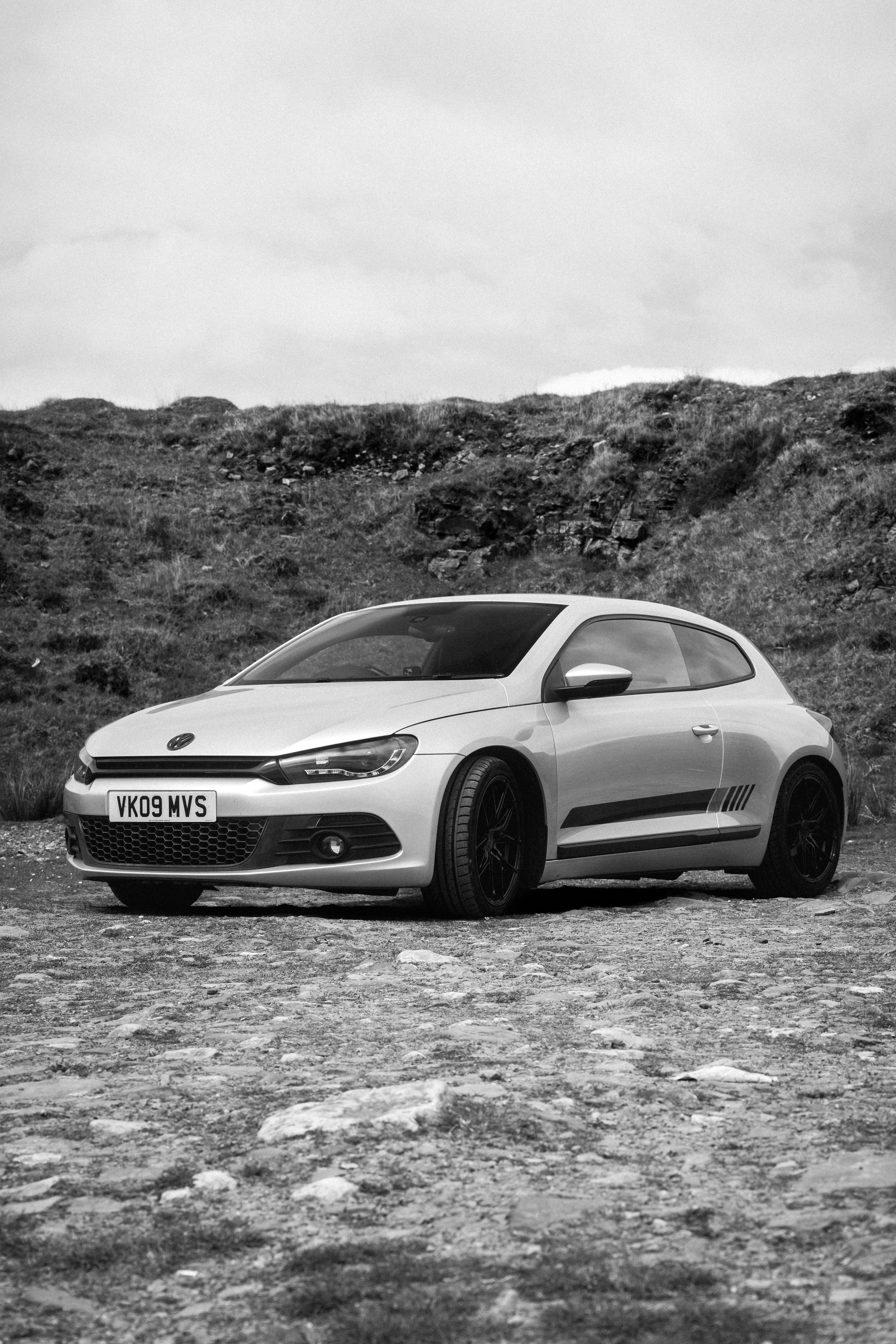 A silver hatchback car parked on a gravel surface with a hill in the background, black and white photo.