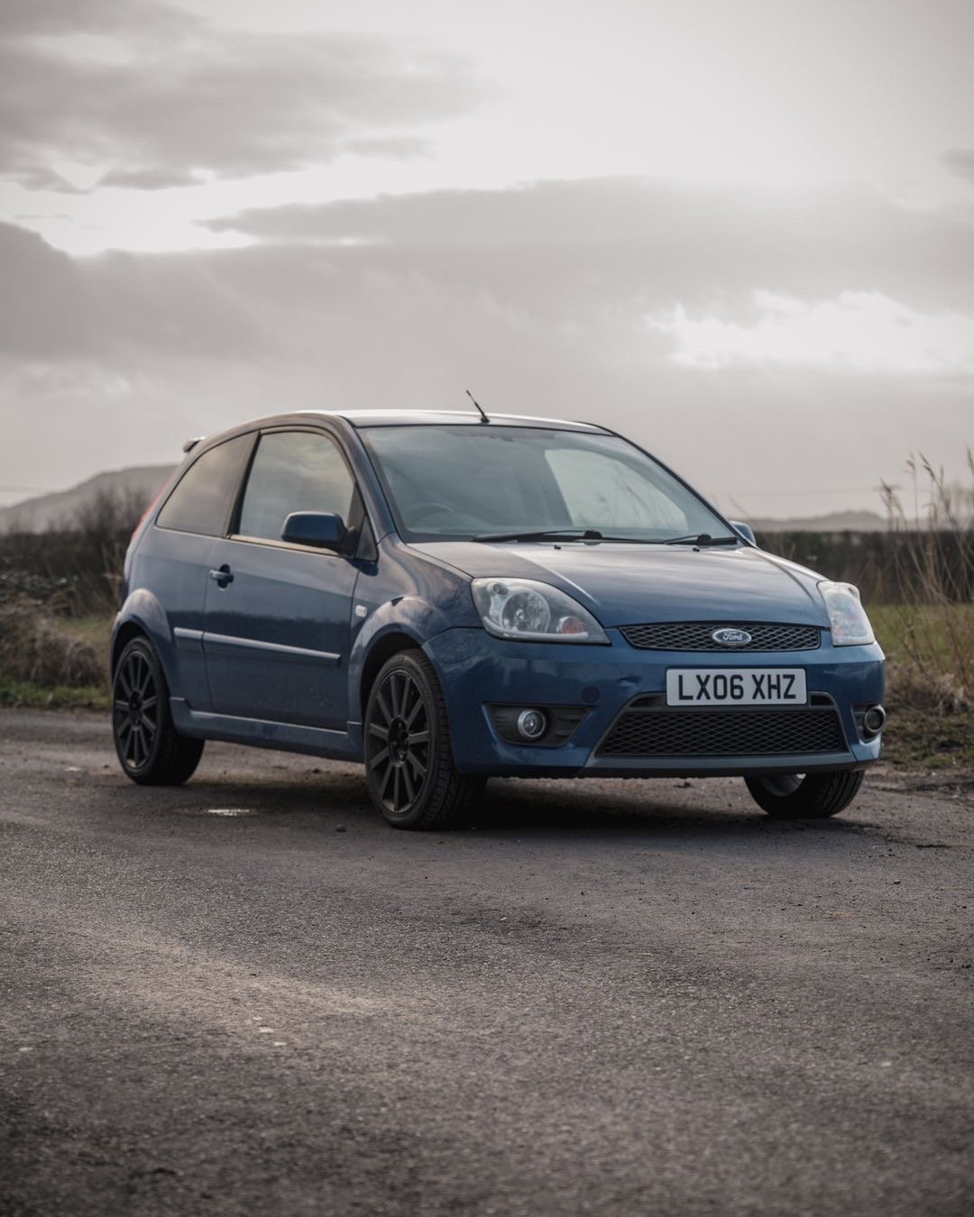 A blue Ford Fiesta parked on the side of a rural road with a cloudy sky in the background.