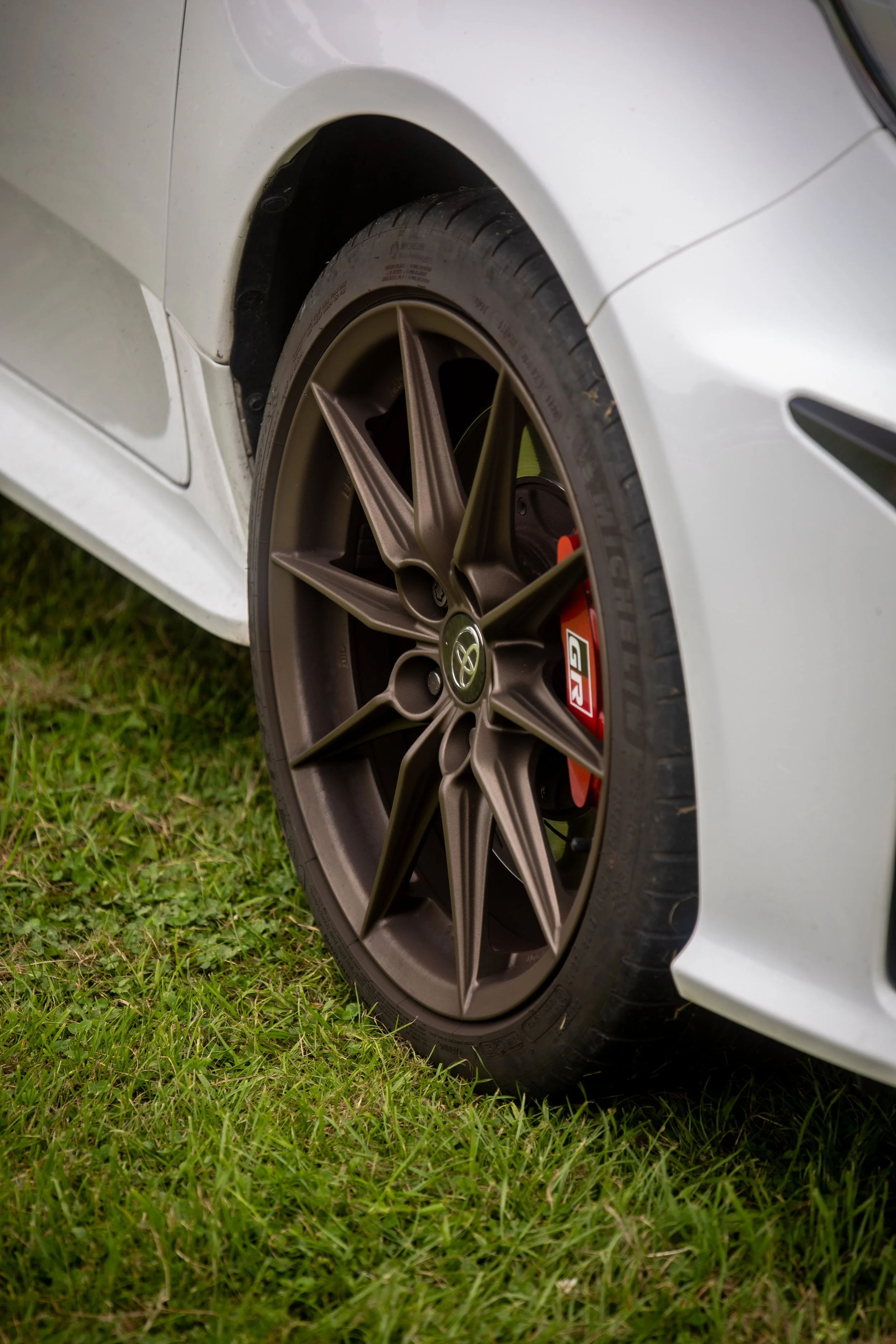 Close-up of a car's front tire and wheel on grass, featuring a Toyota logo, BRZ brake caliper, and a white body panel.