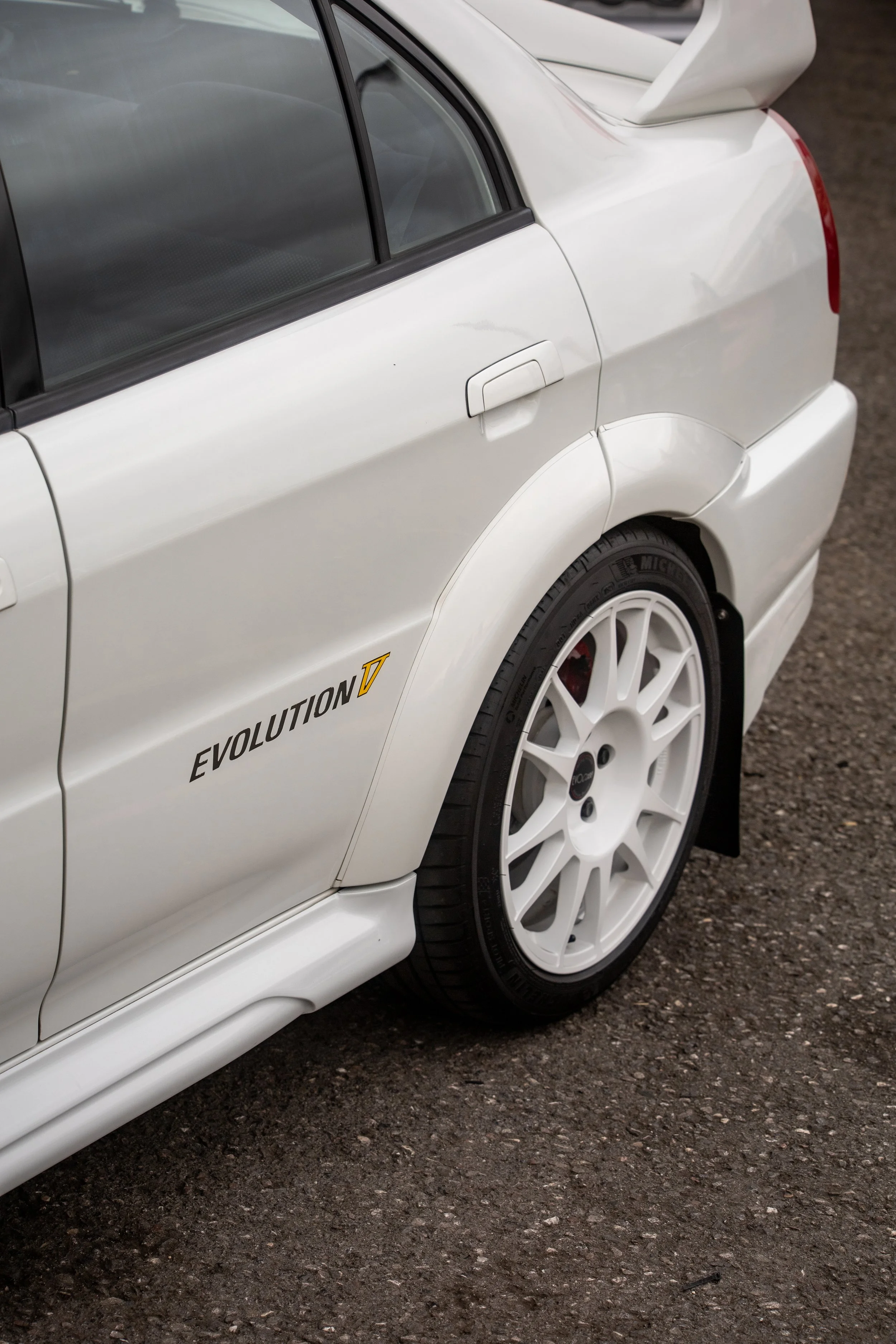 Close-up of a white rally car with an 'Evolution VII' sticker, showing the rear wheel, part of the rear quarter panel, and side window on a gravel surface.