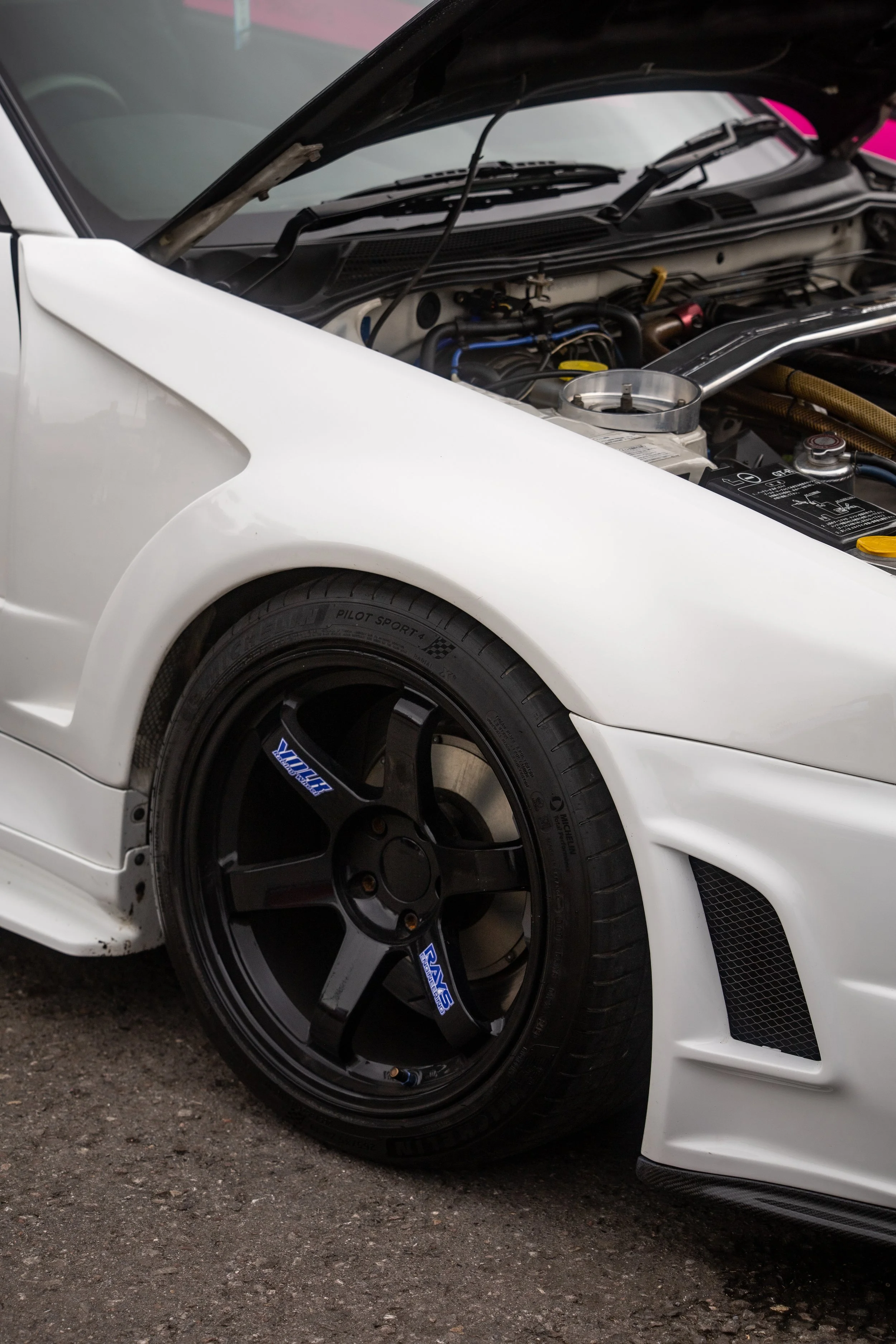Close-up of a white sports car with black racing wheels, front fender, and open hood revealing engine components.