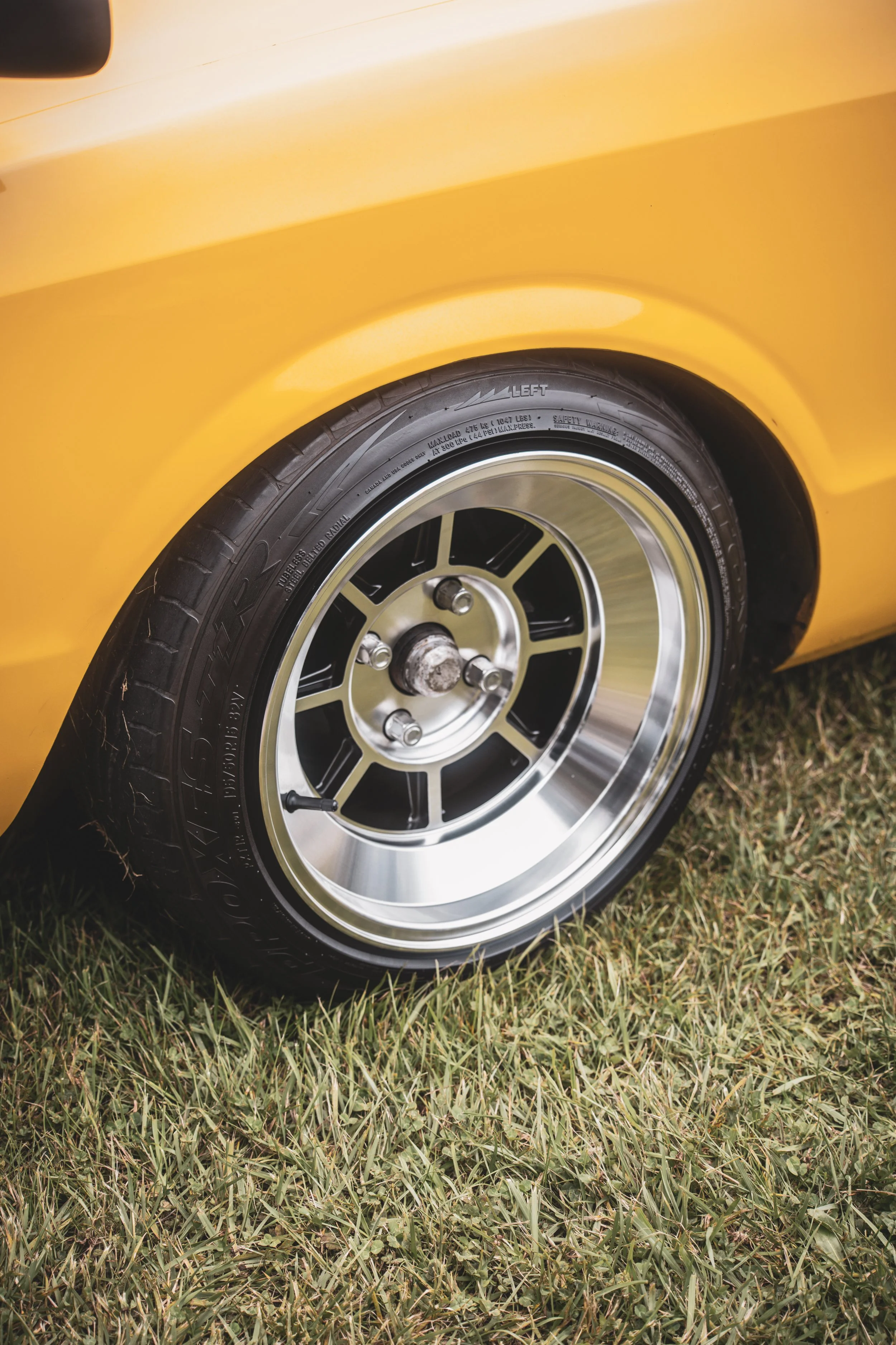 Close-up of a car's rear wheel, featuring a chrome rim and black tire, on grass with a yellow vehicle body.