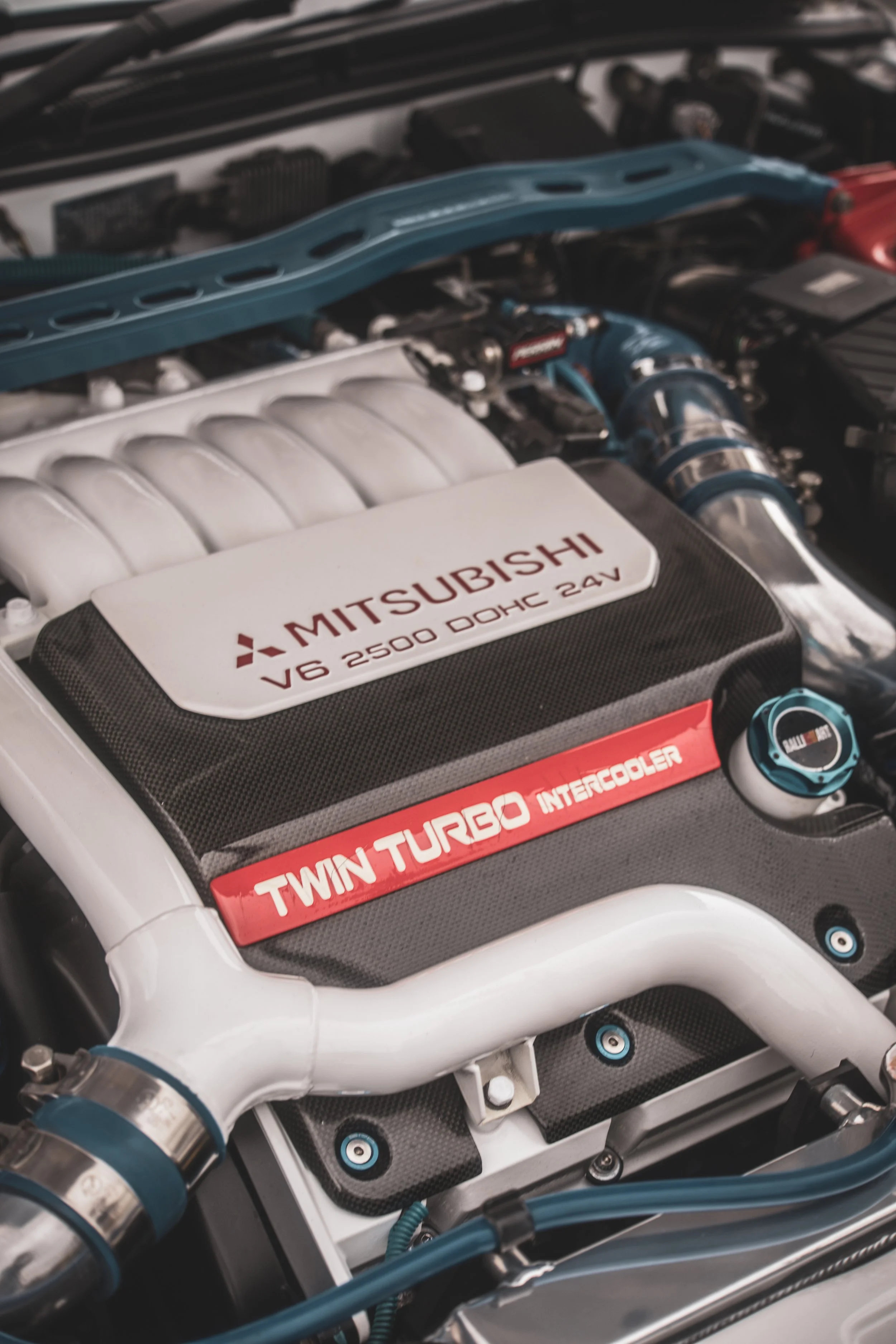 Close-up of a Mitsubishi car engine showing a black cover labeled "V8 2500 DOHC 24V" and "Twin Turbo Intercooler" with visible metal and plastic engine components.