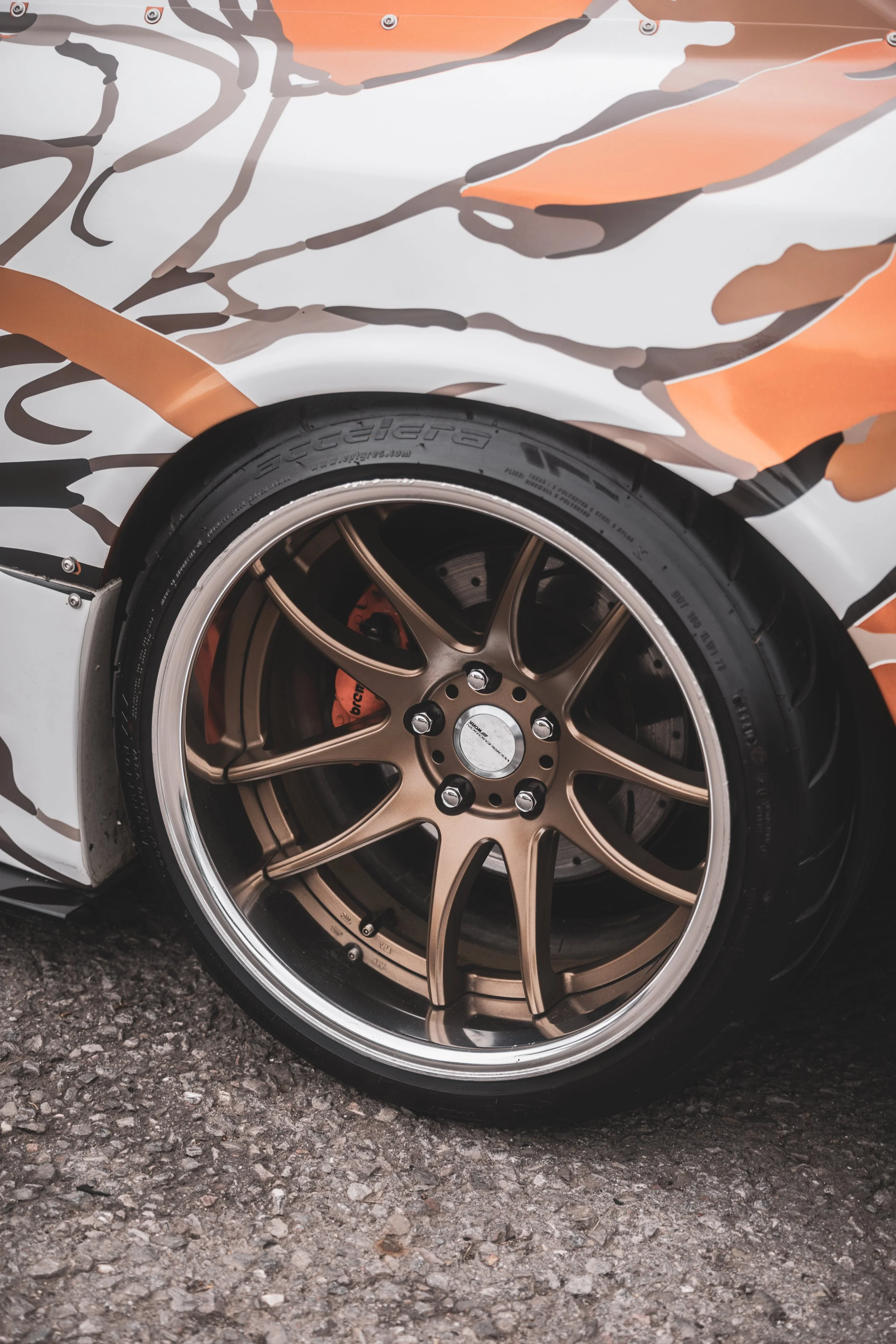 Close-up of a car's gold alloy wheel, black Tire, orange brake caliper, and custom camouflage paint job on the car body.