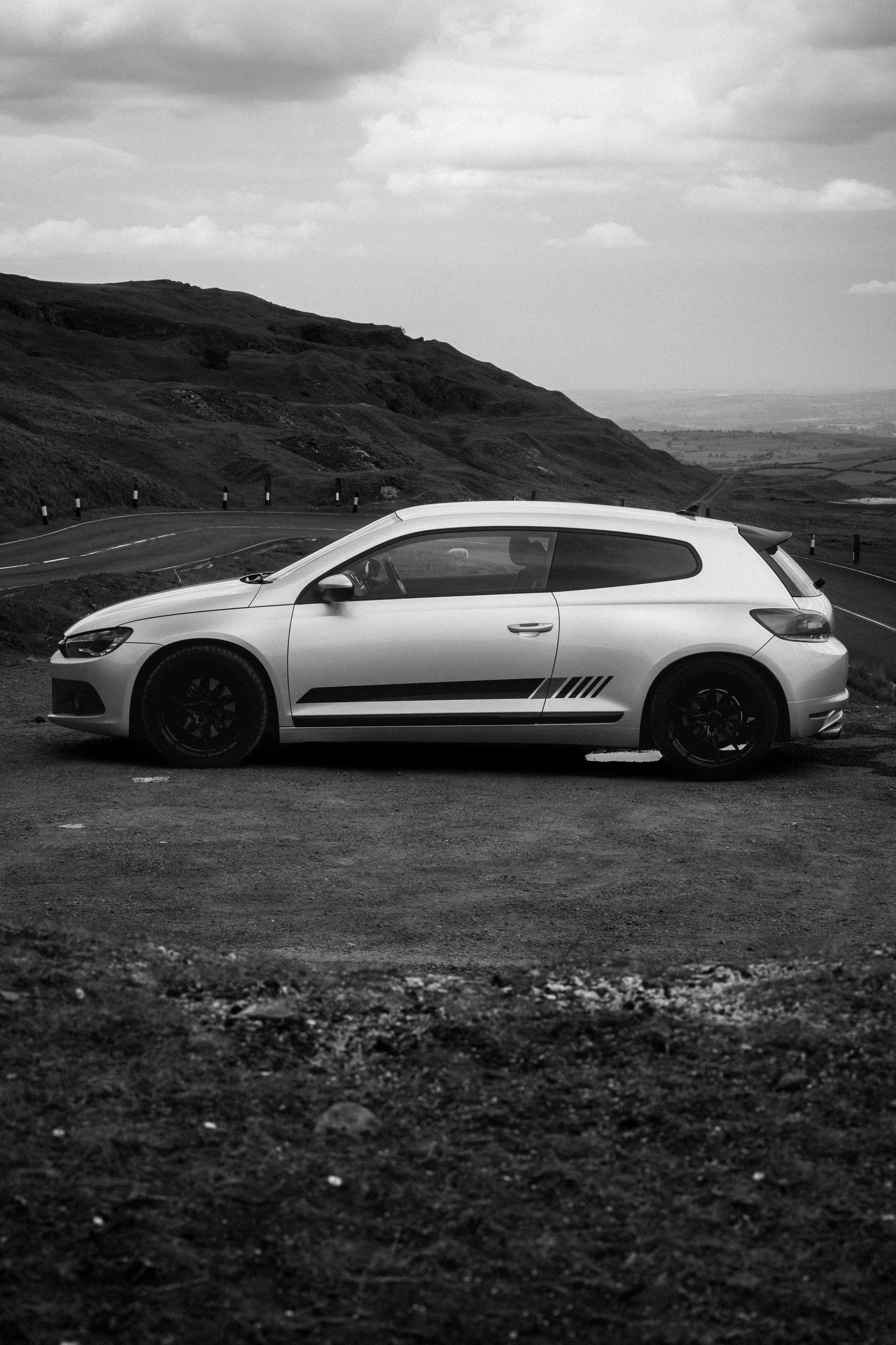 A silver hatchback car parked on a dirt area along a winding rural road with hills and cloudy sky in the background.