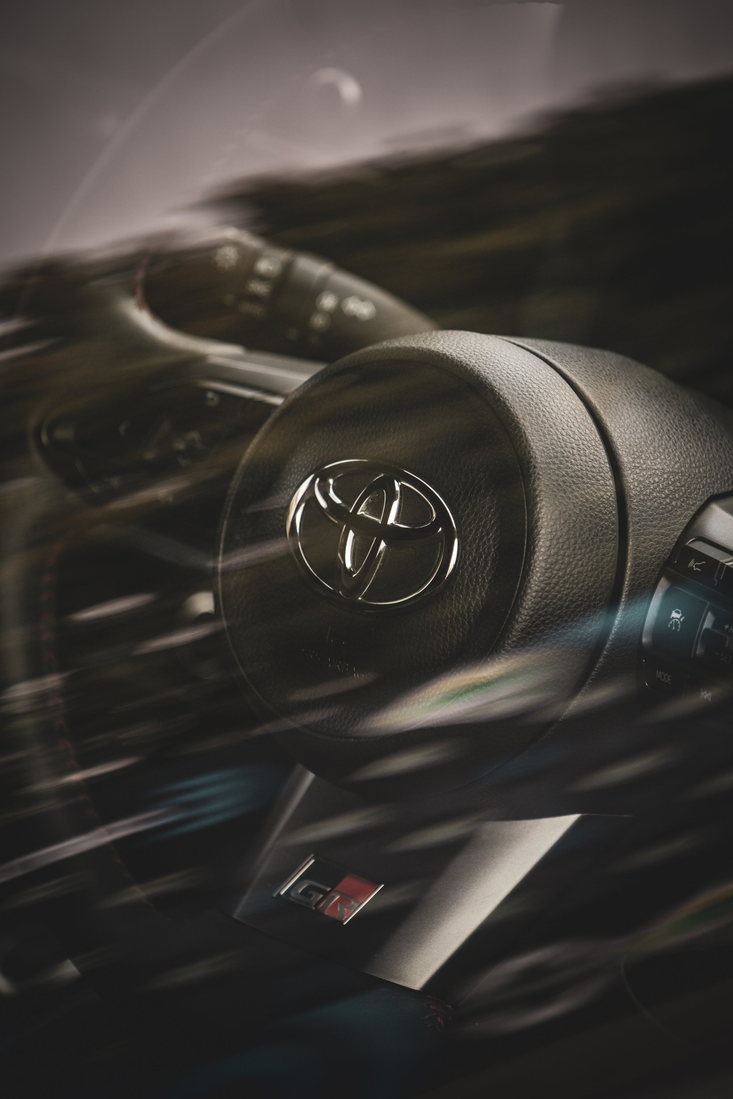 Close-up of a Toyota car steering wheel with the Toyota logo, and a GT badge below, with motion blur streaks across the image.