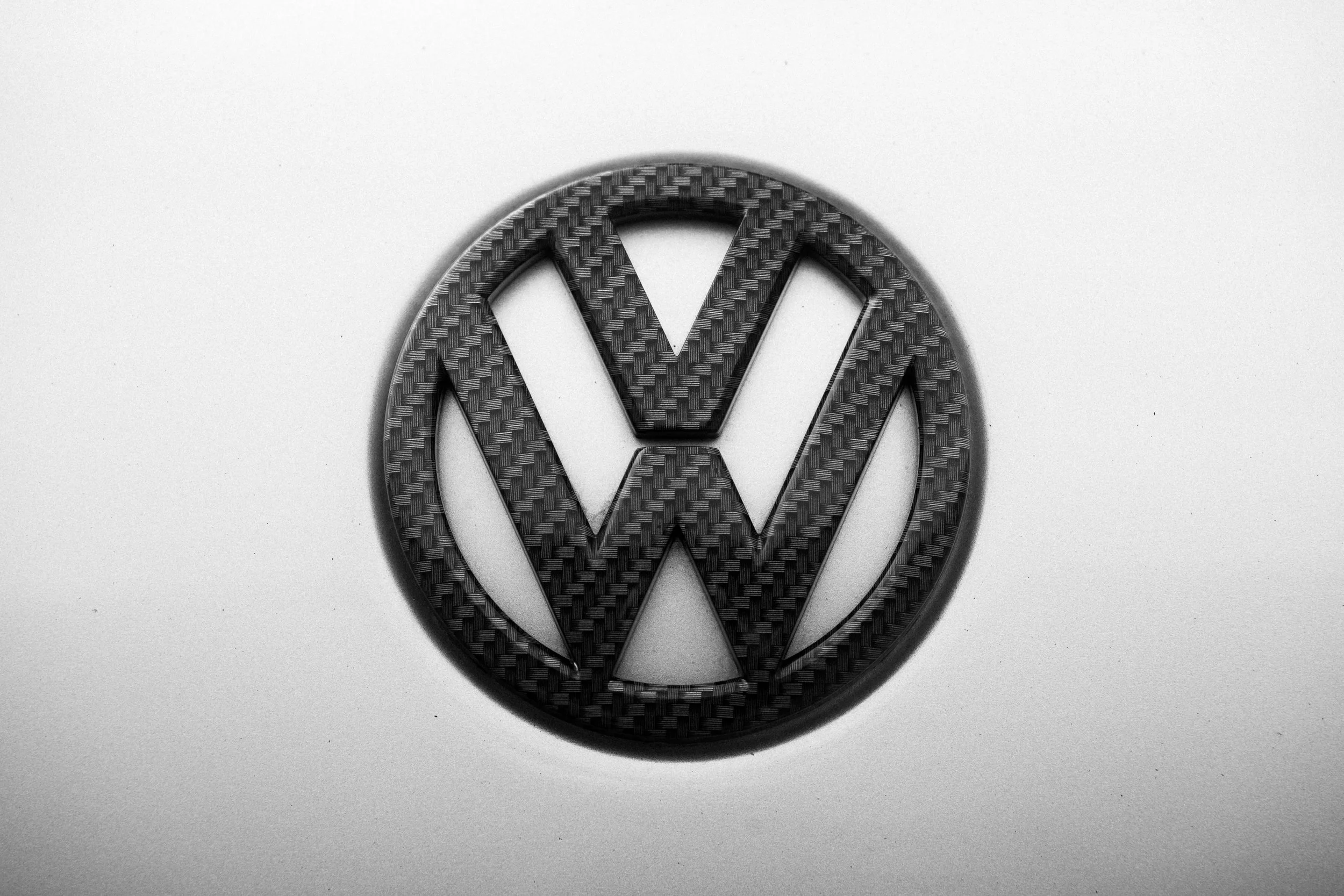 A close-up of a Volkswagen logo with a textured carbon fiber pattern on a silver metallic surface.