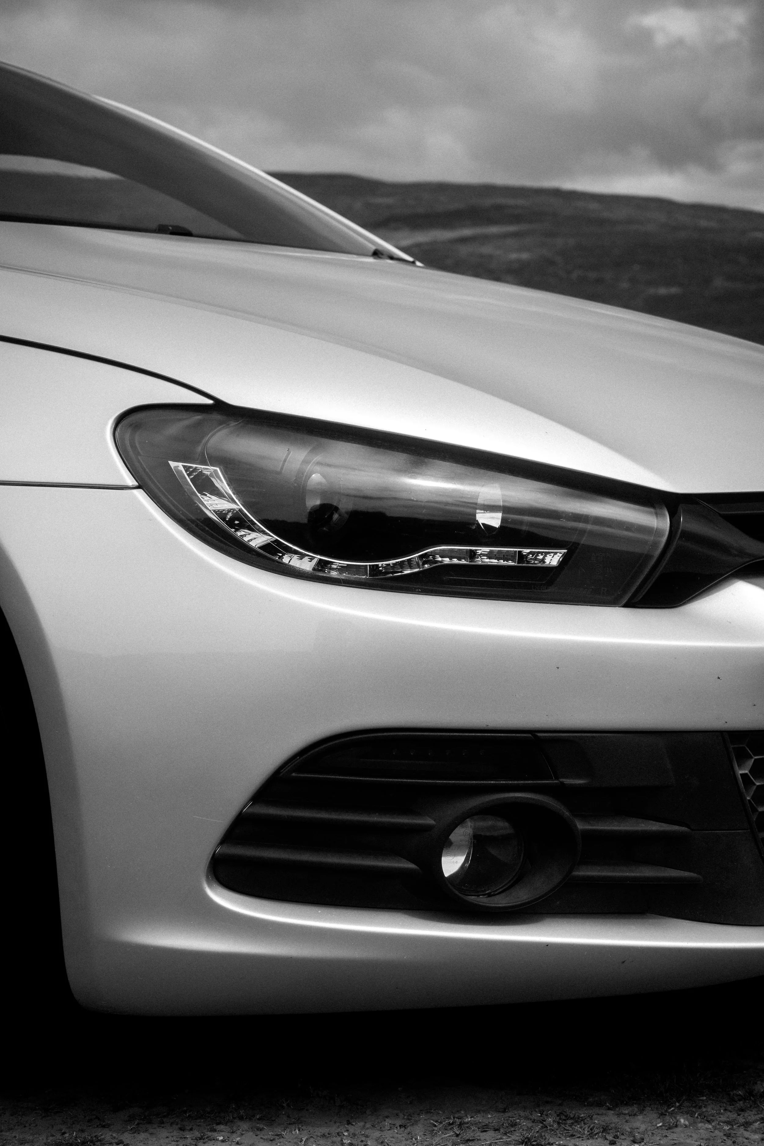 Close-up of the front partial view of a white modern car, showing the headlight and fog light area, against a background of cloudy sky and rolling hills.