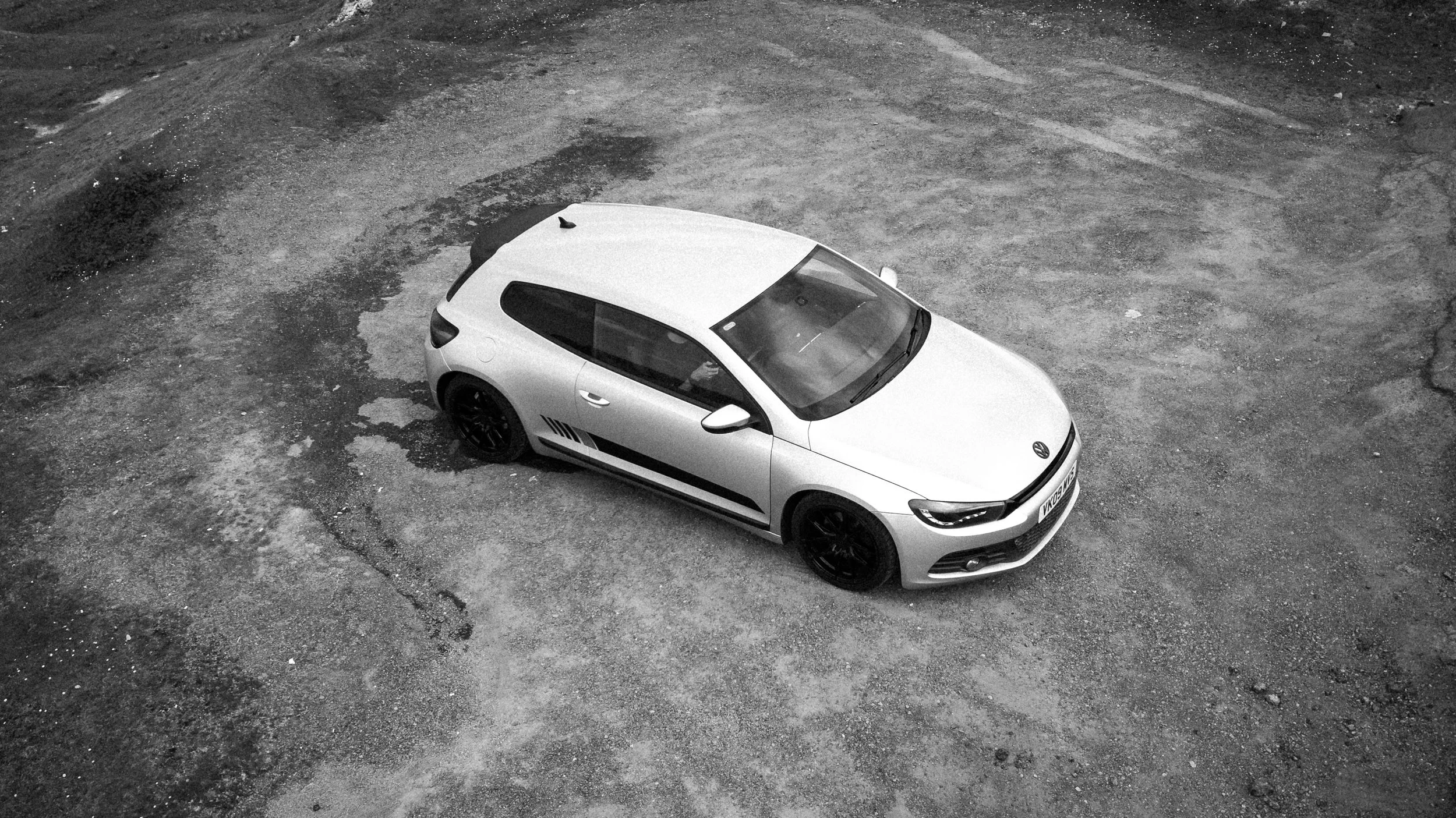 A white VW car parked on a gravel surface, viewed from above in black and white.