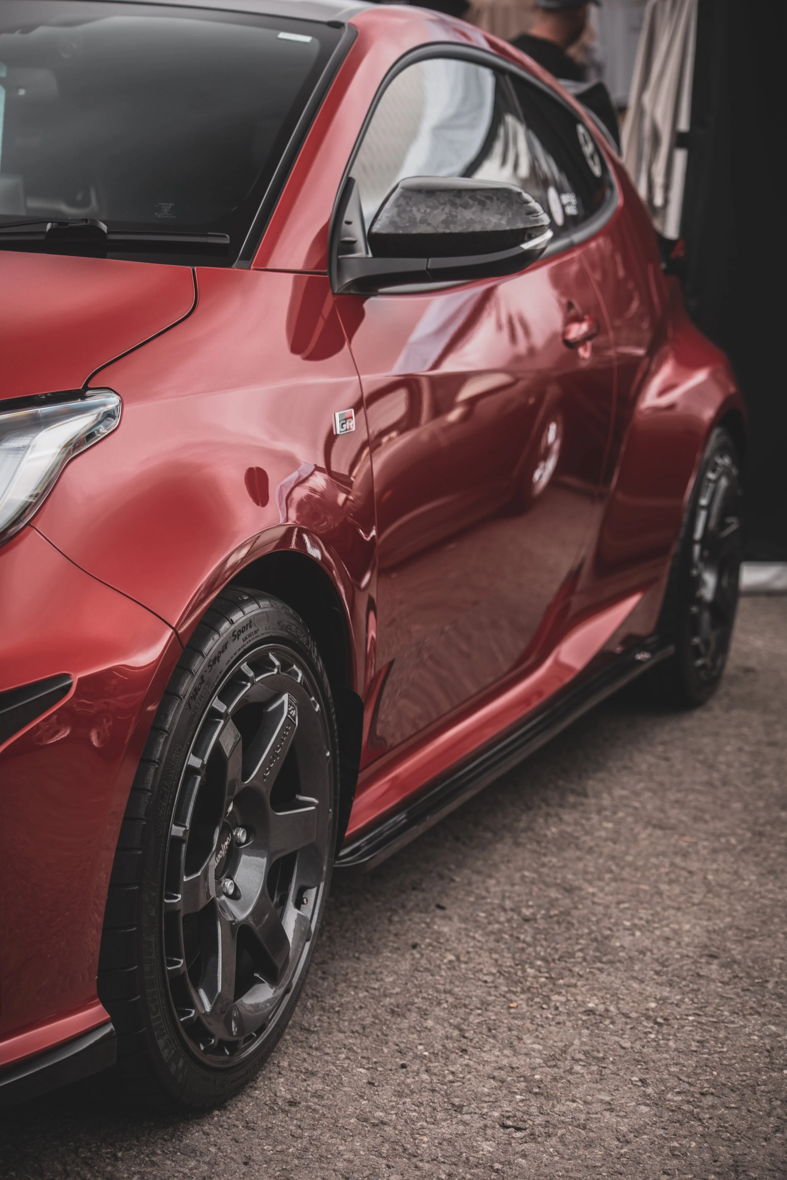 Close-up of a red sports car with black wheels, showing the front and side, parked on a paved surface with a person in the background.