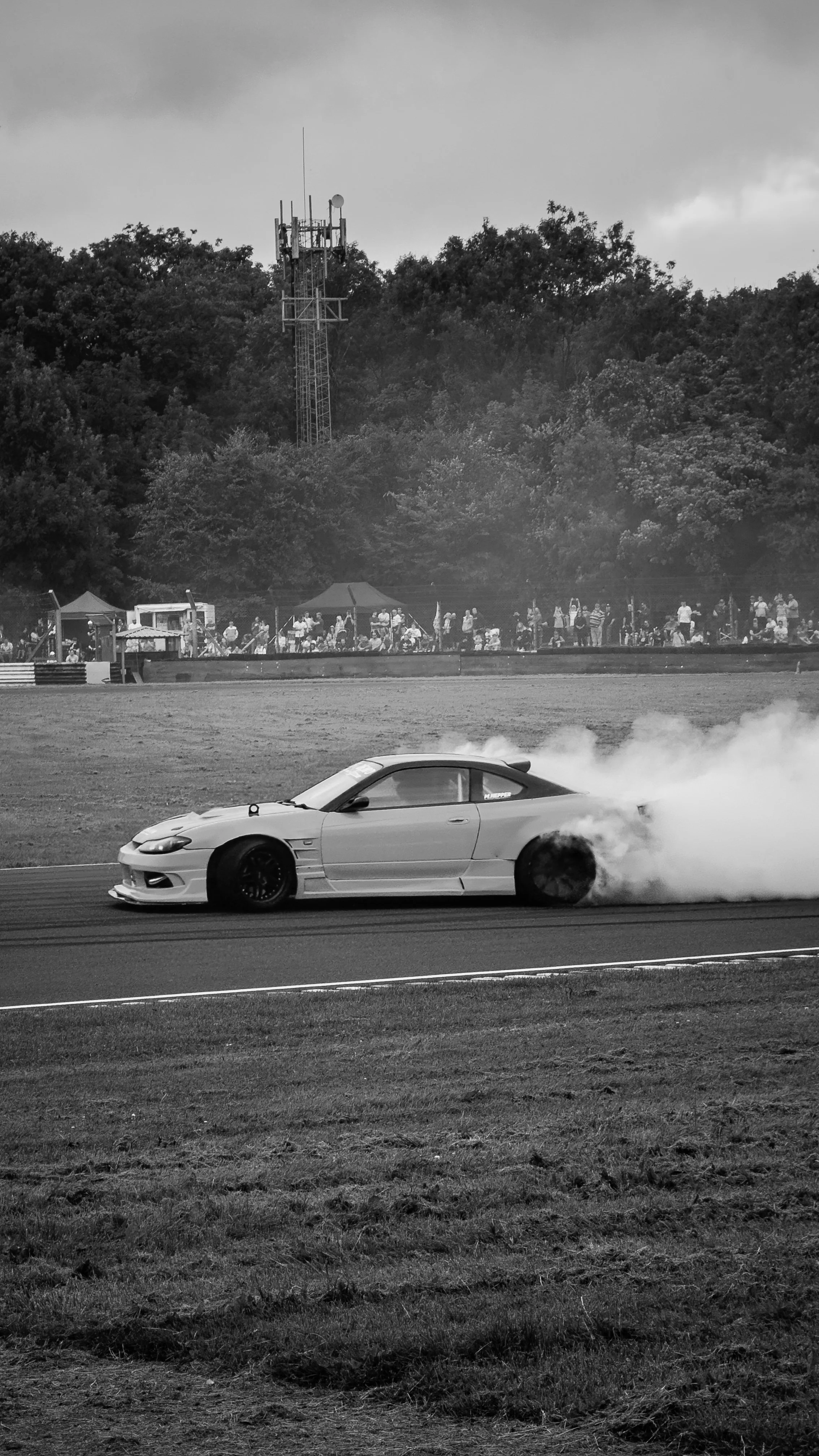 A race car drifting on a track producing smoke from its rear tires during a race event with spectators in the background and a wooded area with a communication tower beyond.