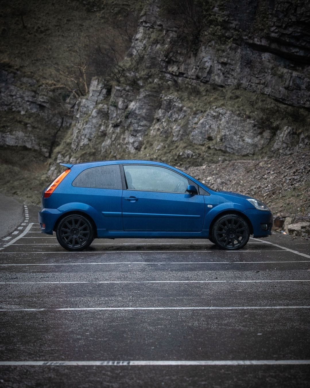 A blue hatchback car parked in a parking lot next to a rocky hillside at dusk.