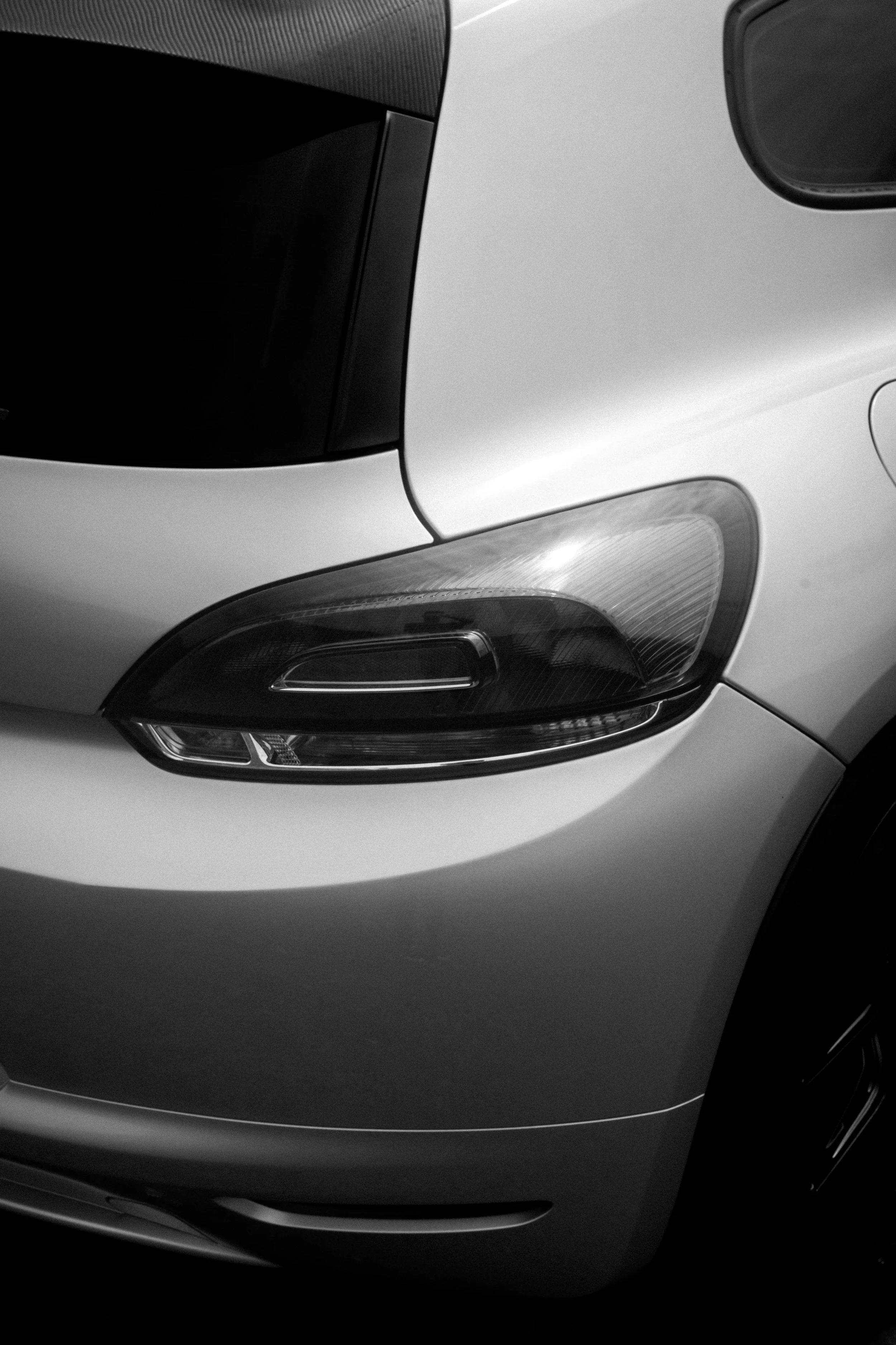 Close-up of the front left part of a silver car, highlighting the headlight and part of the hood, in black and white.