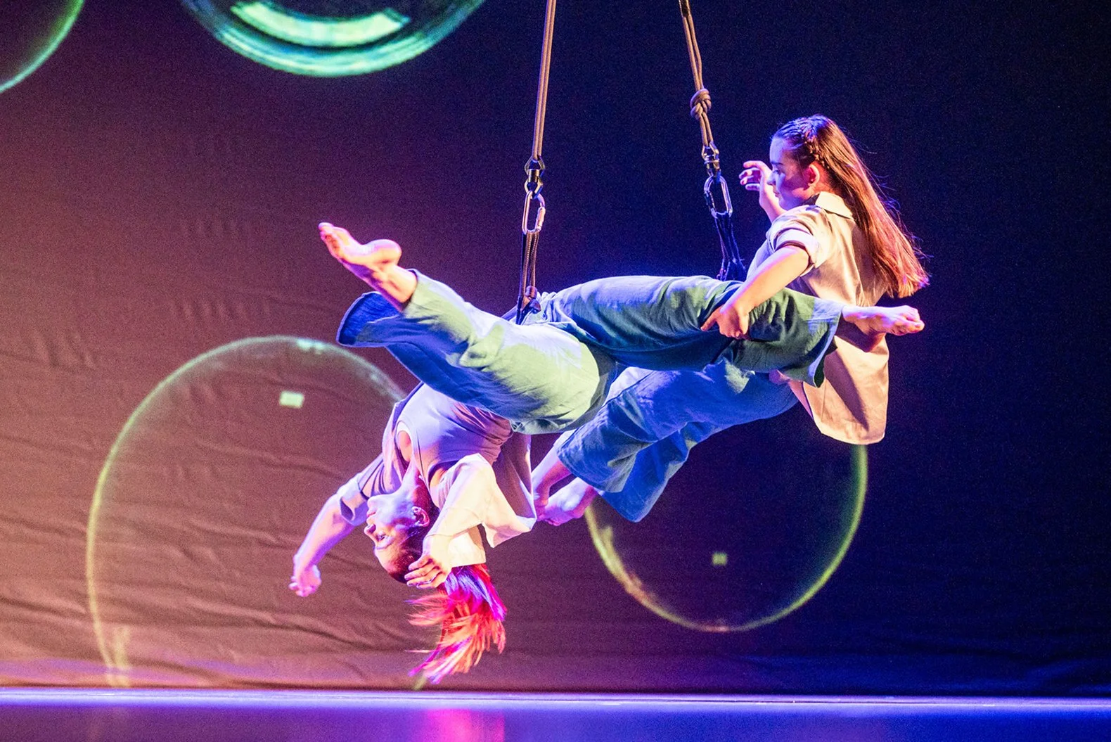 zwei Akrobatinnen hängen an Seilen auf der Bühne vor einem Hintergrund mit großen Blasen