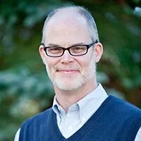 Portrait of a middle-aged man with glasses, short gray hair, wearing a blue sweater over a collared shirt, standing outdoors with greenery in the background.