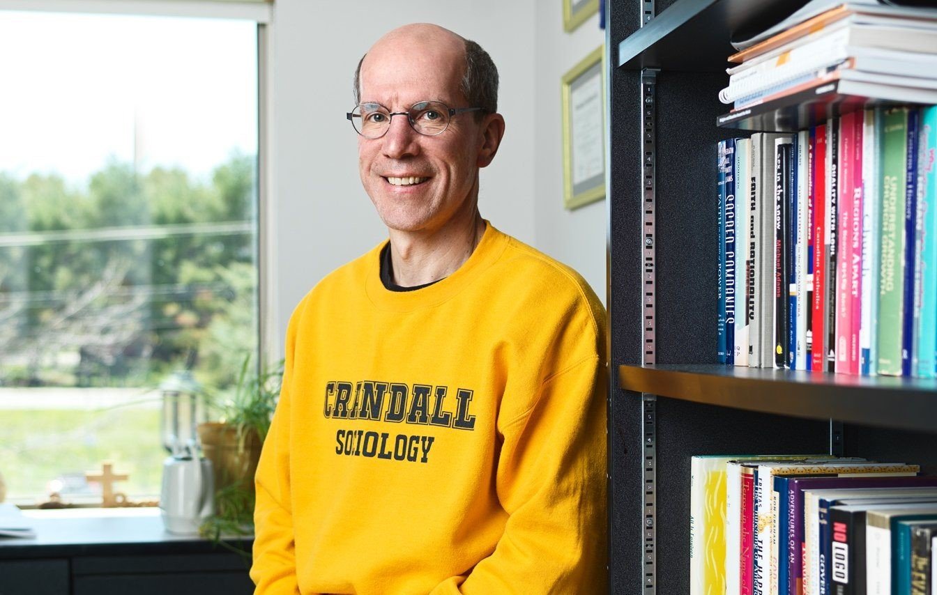 A smiling man in glasses and a yellow sweatshirt with 'Crandall Sociology' stands indoors near a bookshelf filled with books, with a window showing trees and sky outside in the background.