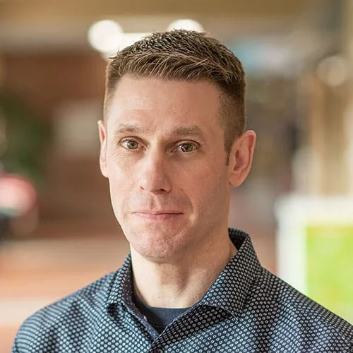 A man with short brown hair and fair skin, wearing a dark patterned shirt, standing outdoors with a blurred background.